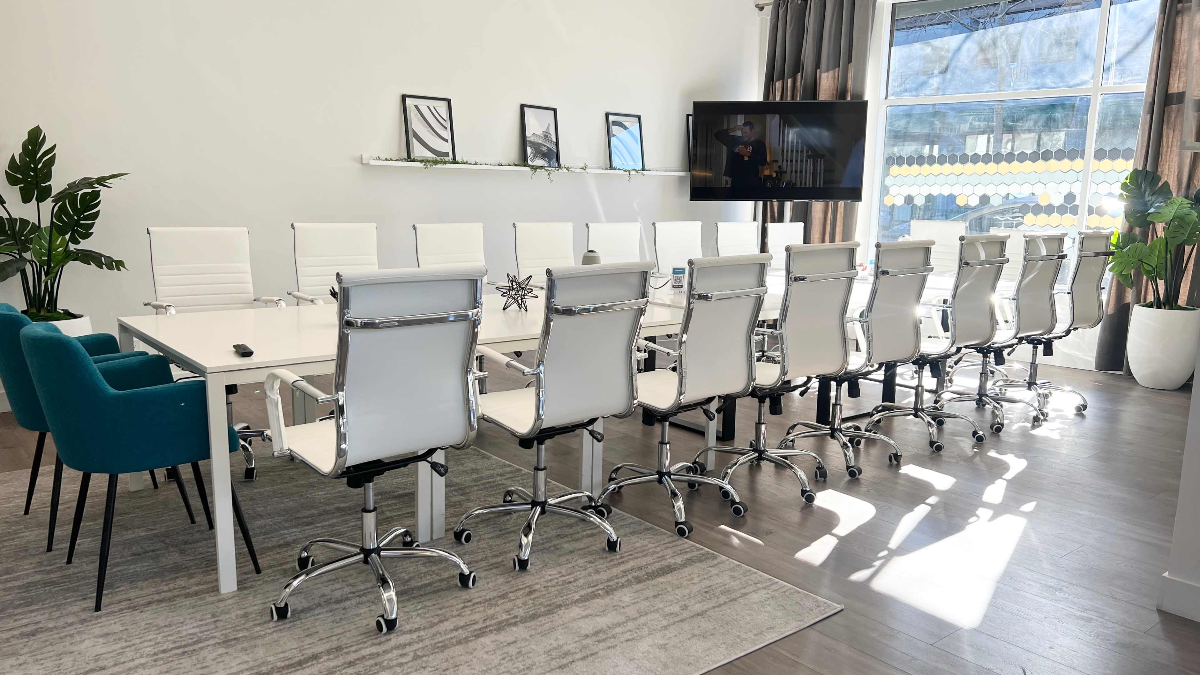 A modern conference room featuring a long rectangular table surrounded by white swivel chairs, with a large window and a television mounted on the wall.
