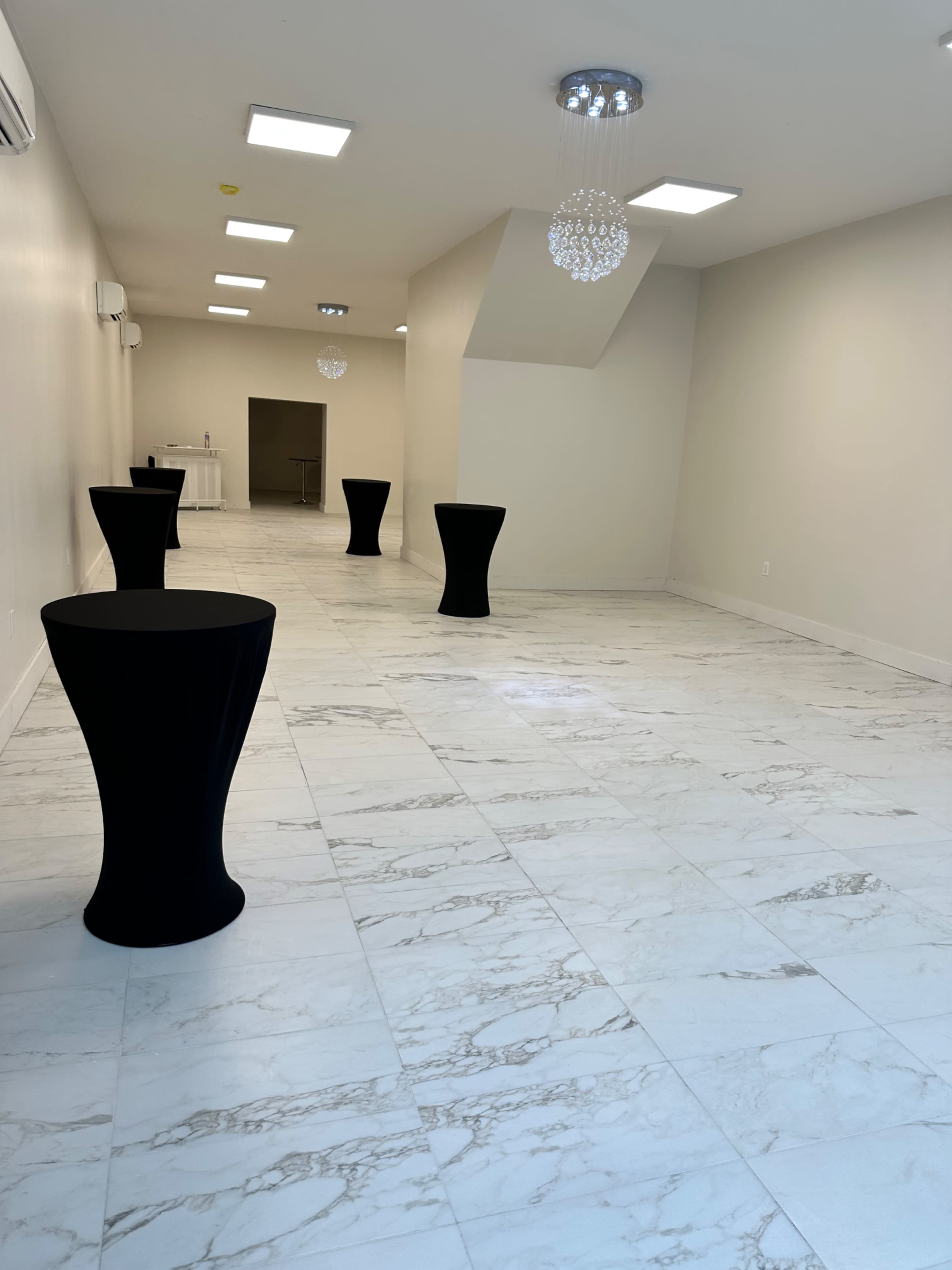 A spacious, minimally decorated event room with marble flooring and several black cocktail tables arranged throughout.
