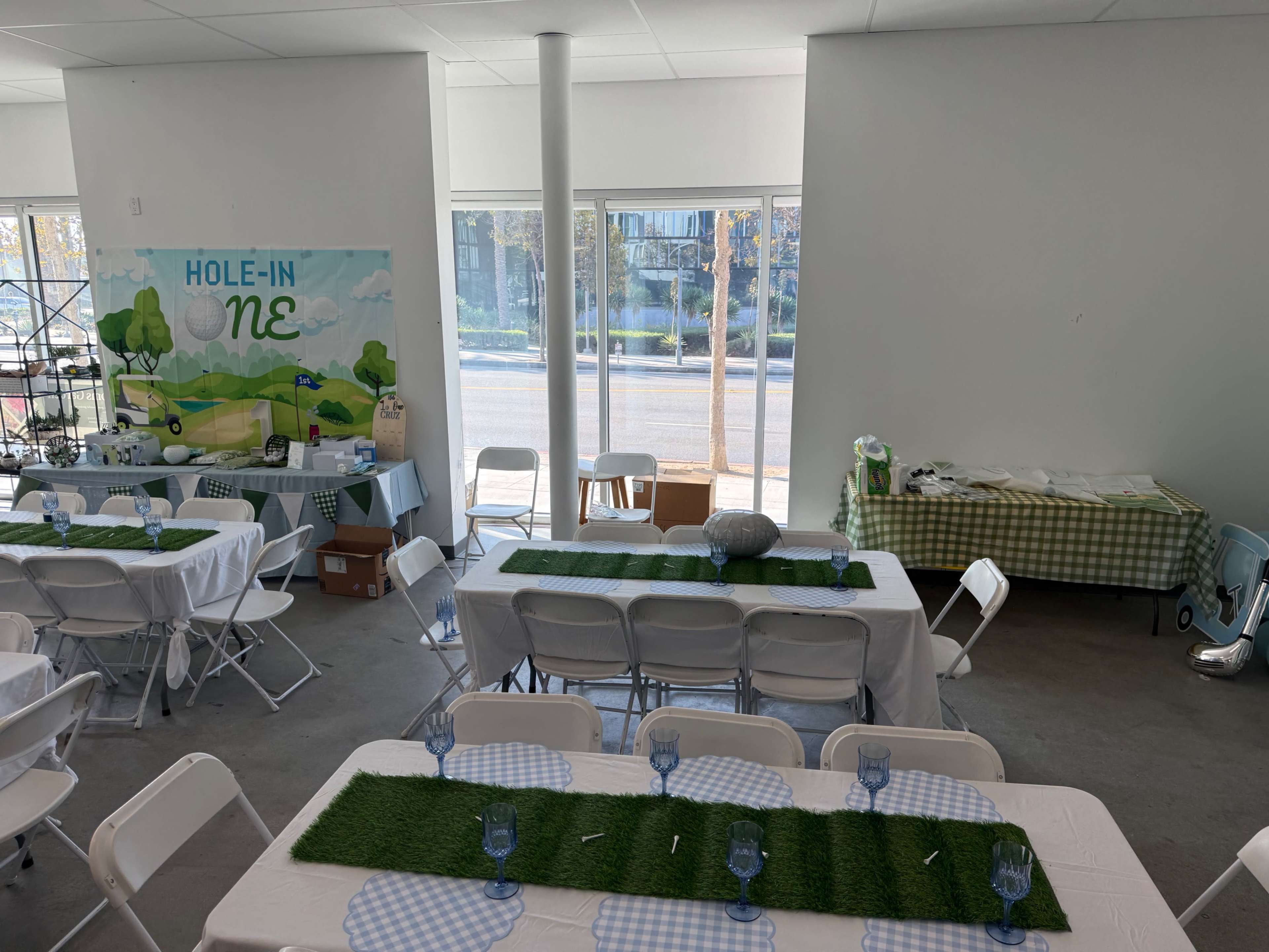 The image shows an indoor event space set up with tables covered in white cloths, decorated with green grass mats and blue glasses, for a gathering themed "Hole-in-One."