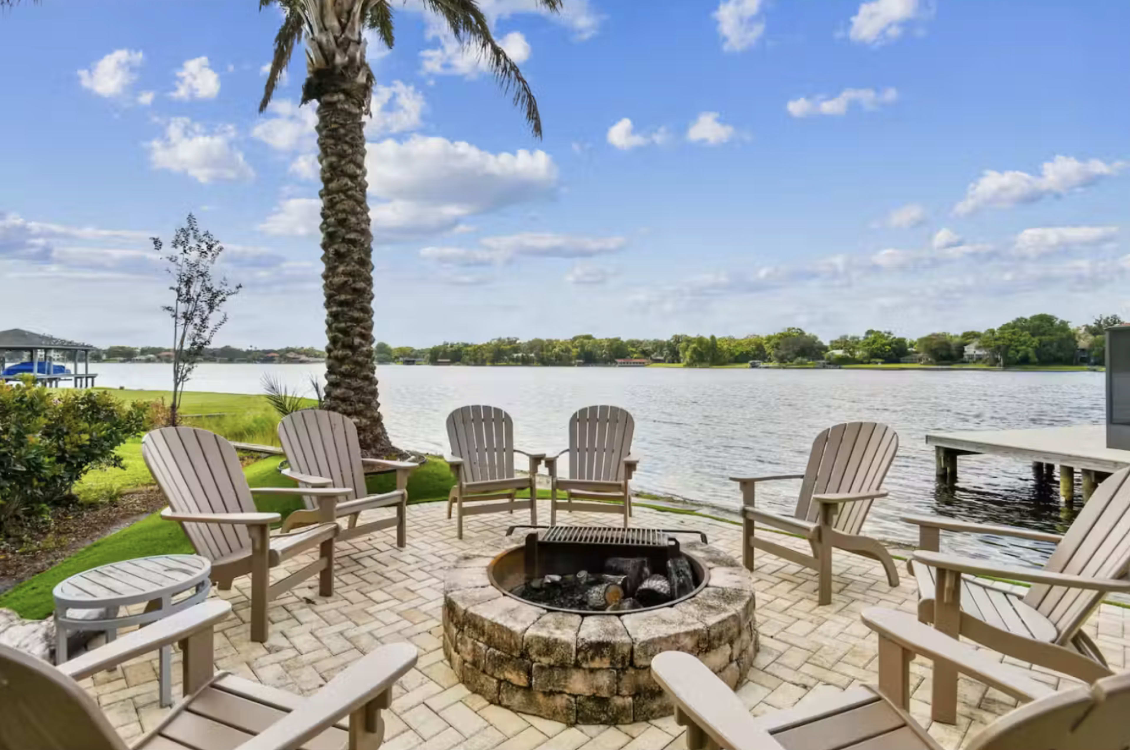 A circular stone firepit surrounded by wooden chairs is situated by a calm lake, with palm trees and a blue sky in the background.