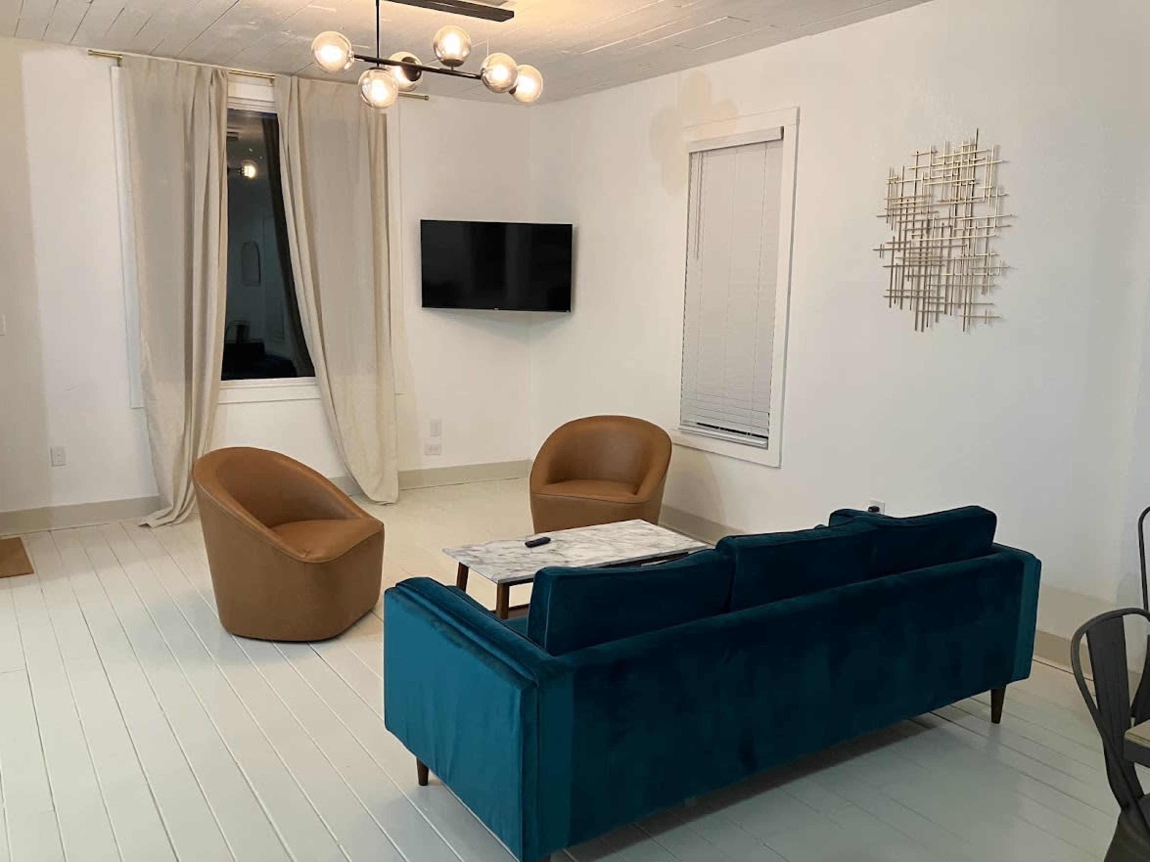 A modern living room with two brown chairs, a teal sofa, a coffee table, a wall-mounted television, and large windows with beige curtains.