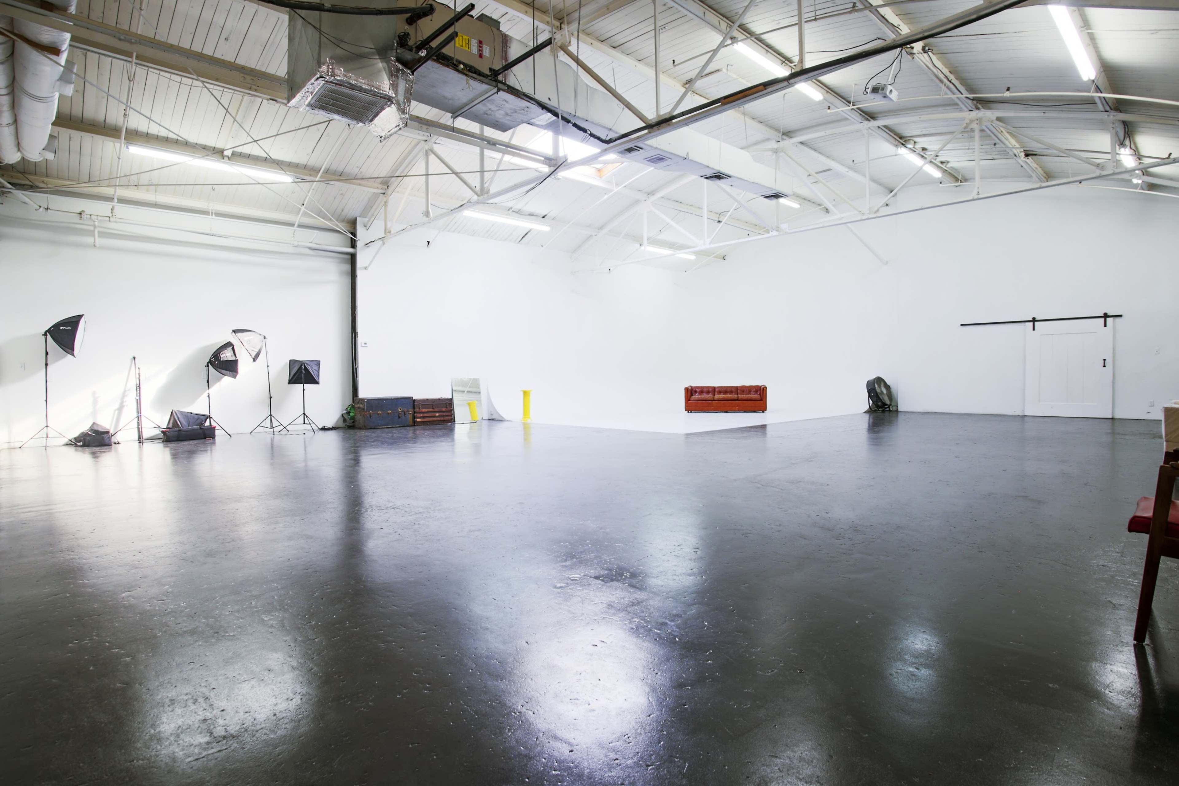 A spacious, empty indoor studio with a polished concrete floor, white walls, and various lighting equipment arranged on one side.