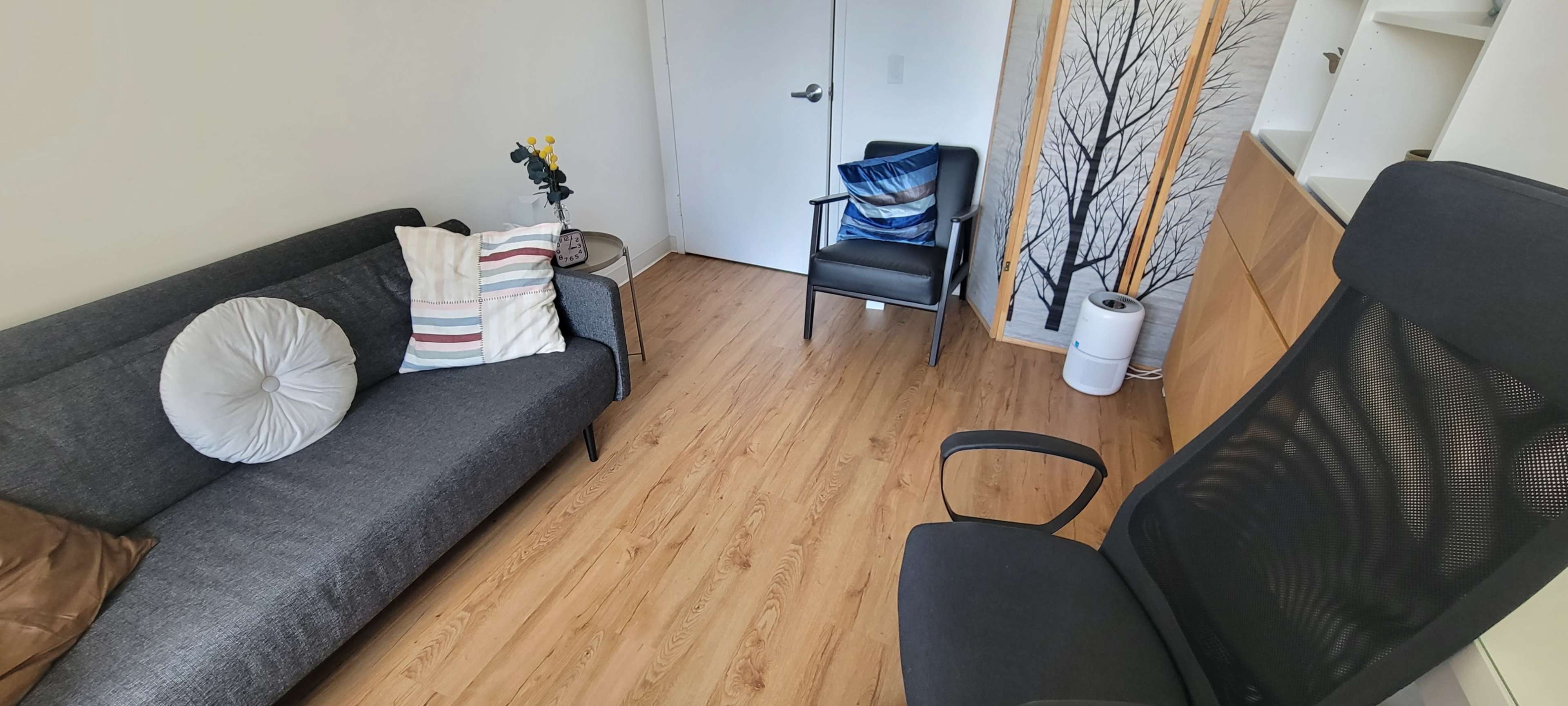 A small living space with a gray sofa, a black chair, and wooden flooring, featuring a door and a decorative wall with a tree design.