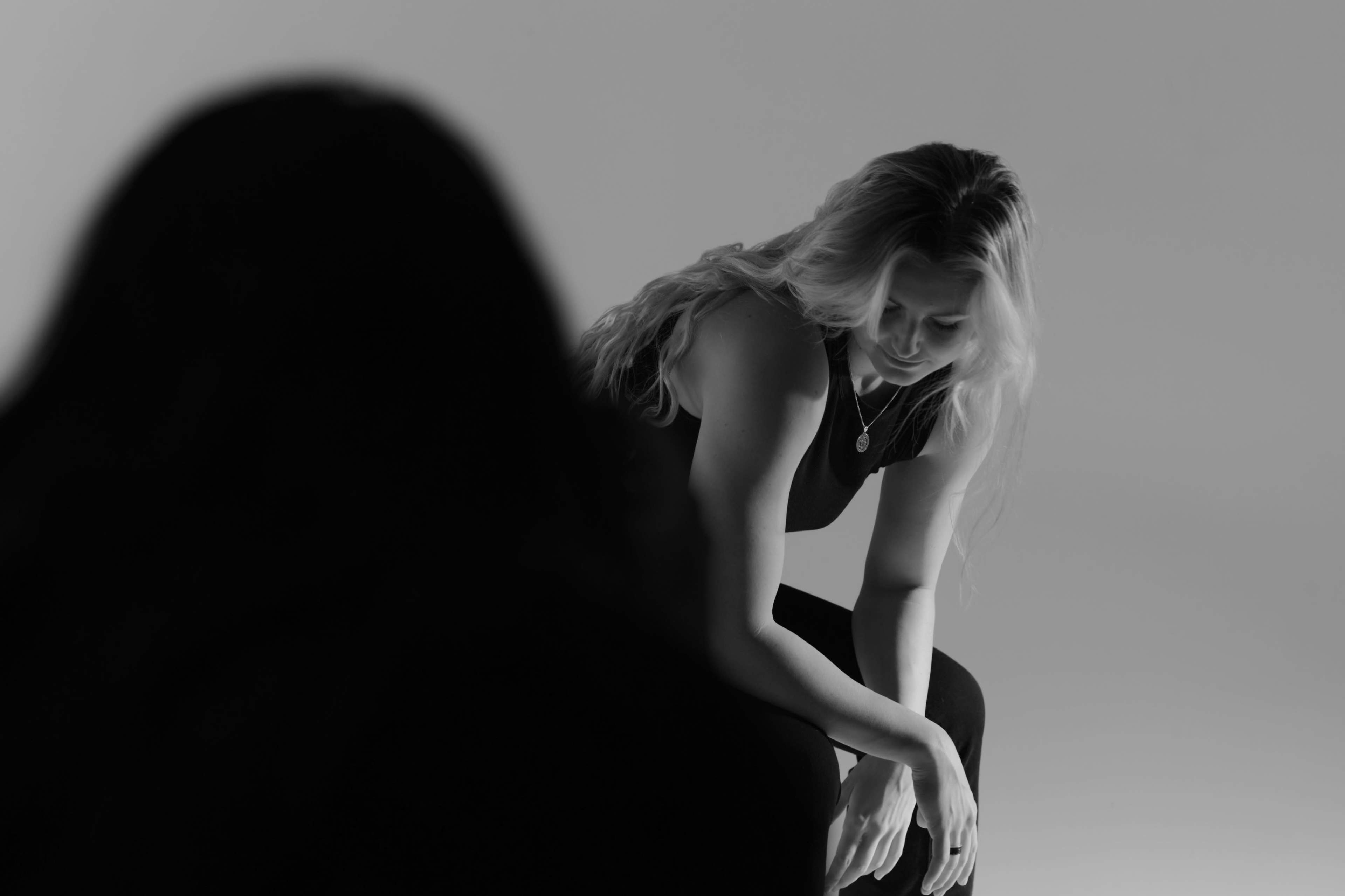 A woman with long blonde hair sits with her head bowed, wearing a dark outfit, while another figure is partially visible in the foreground.