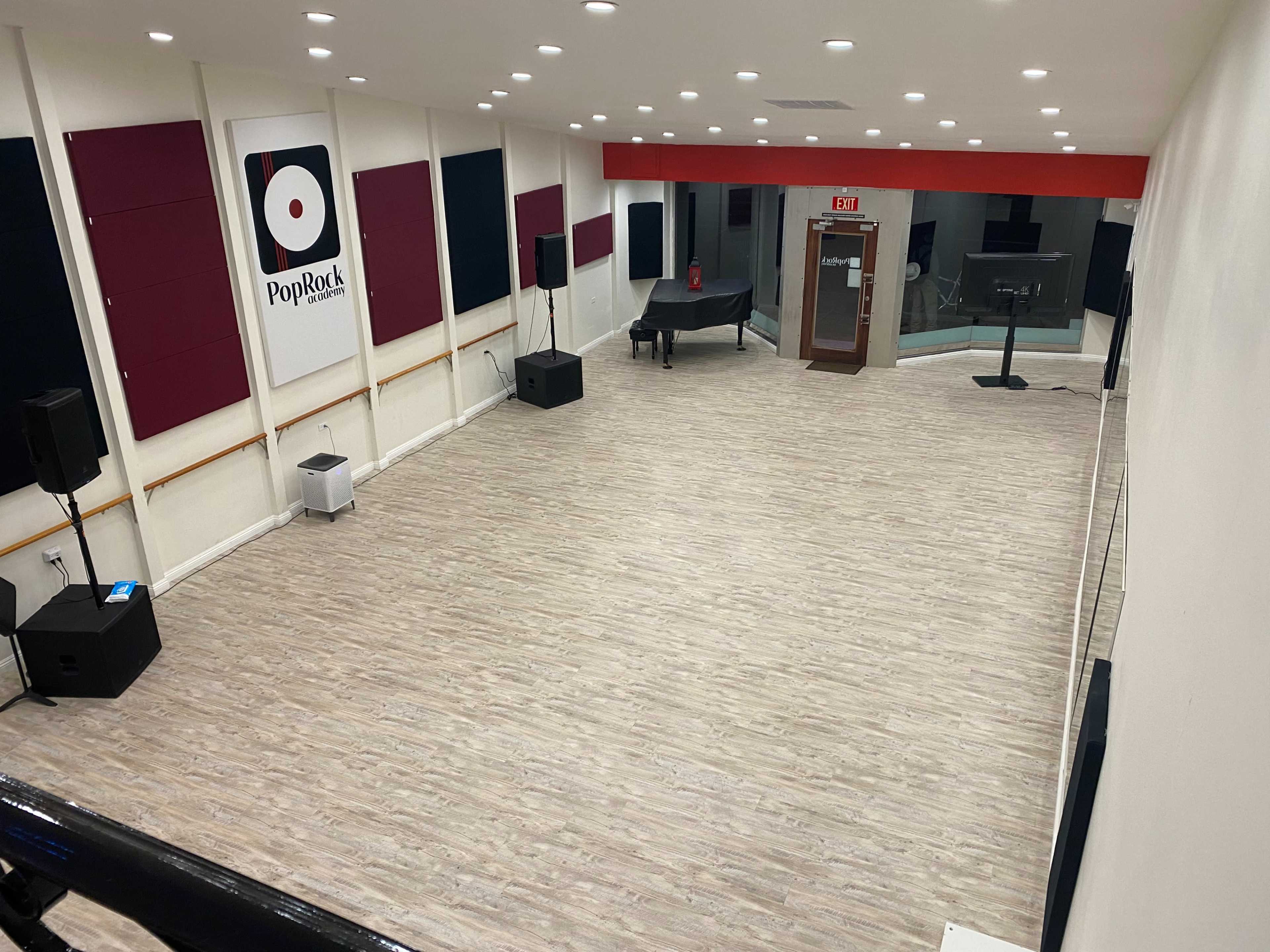 A spacious interior room with a wooden floor, featuring sound equipment, a grand piano, and red and black wall panels, along with a glass entrance.