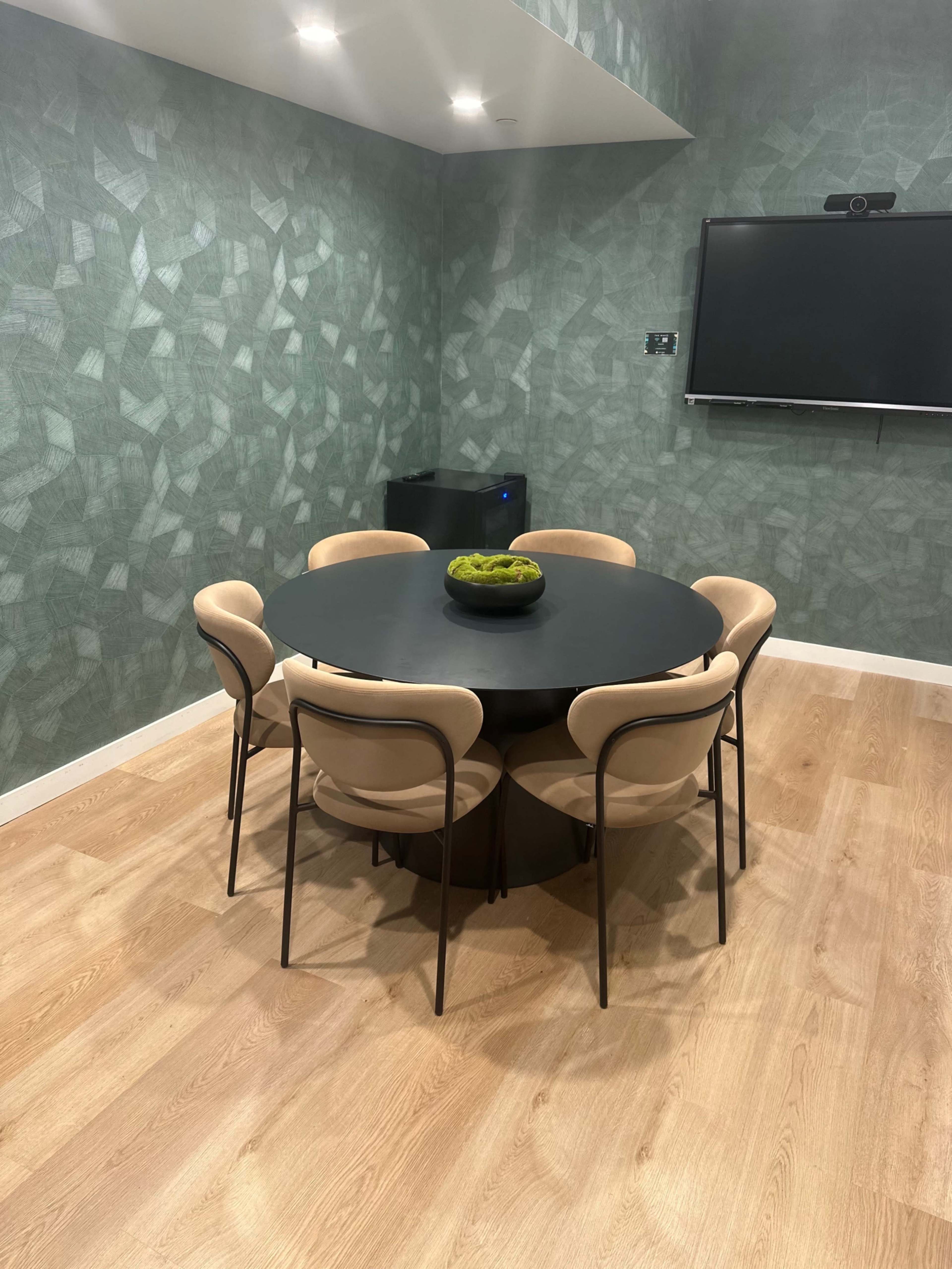 A round black dining table with six chairs is set in a room with green patterned walls and hardwood flooring, featuring a television mounted on the wall.