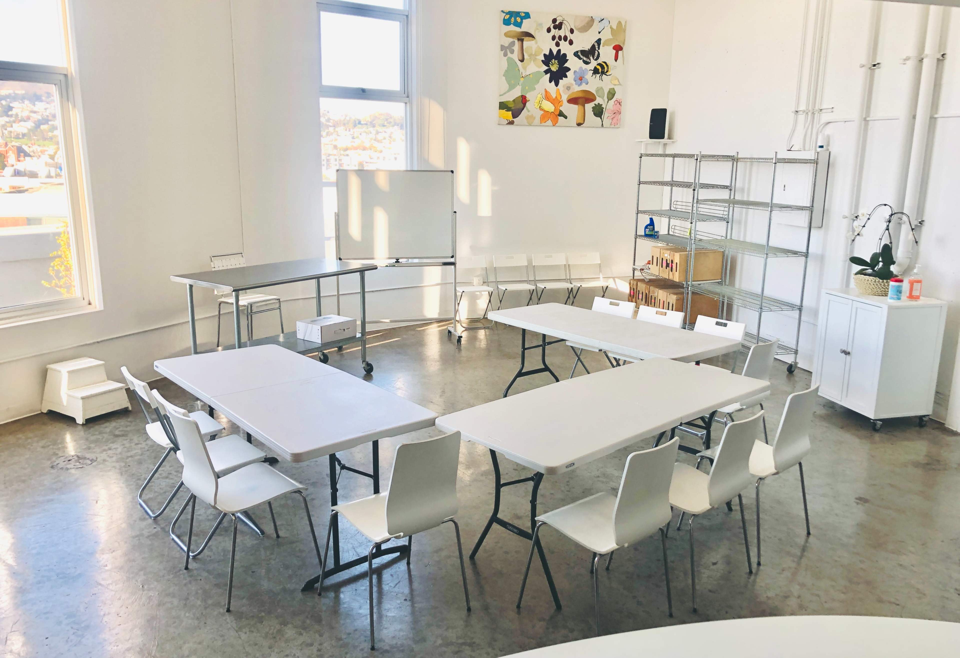A bright, modern classroom with multiple tables arranged in a U-shape and white chairs, situated next to large windows.