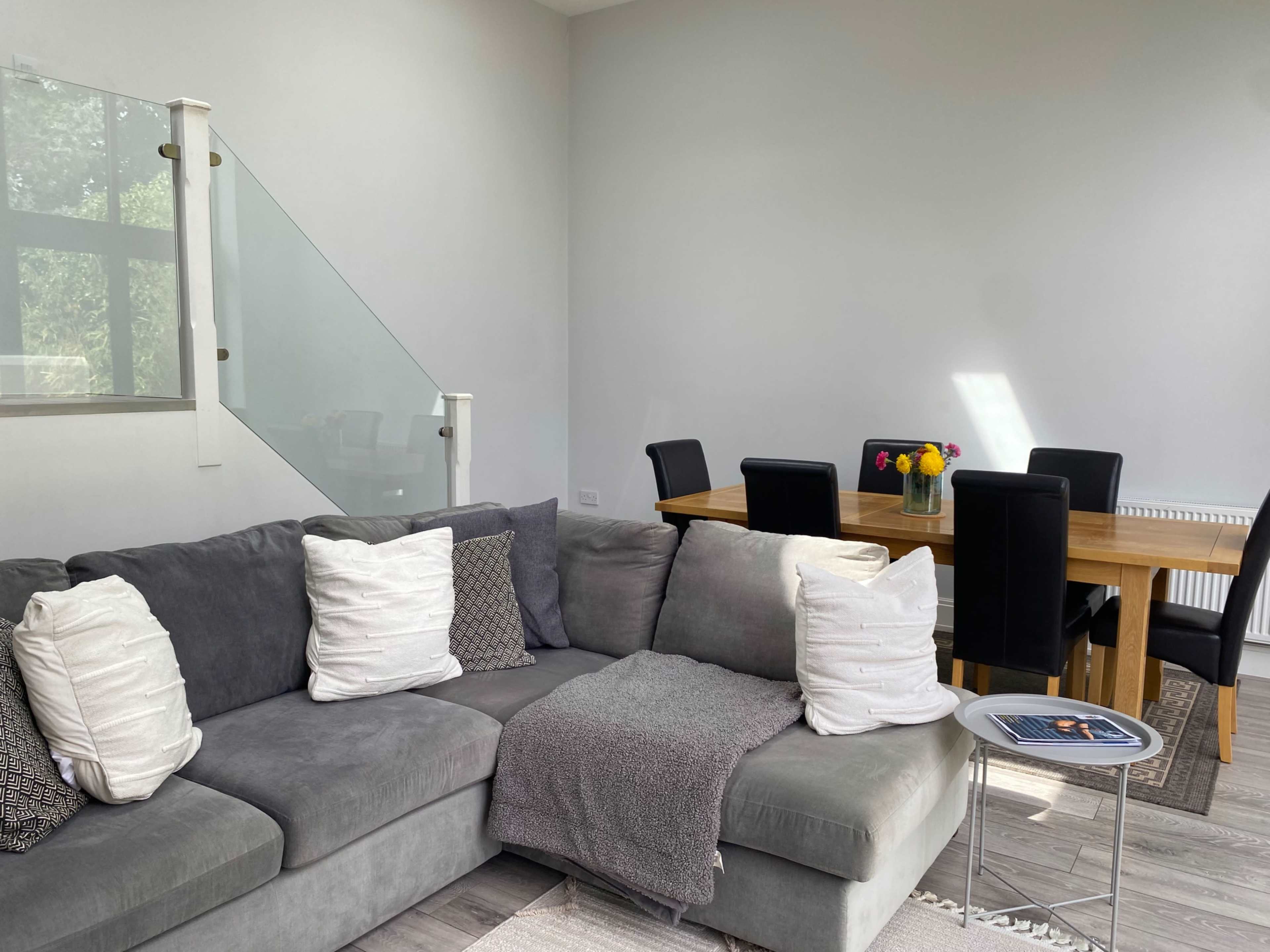 The image shows a modern living area with a gray sectional sofa and a dining table set for six in the background.