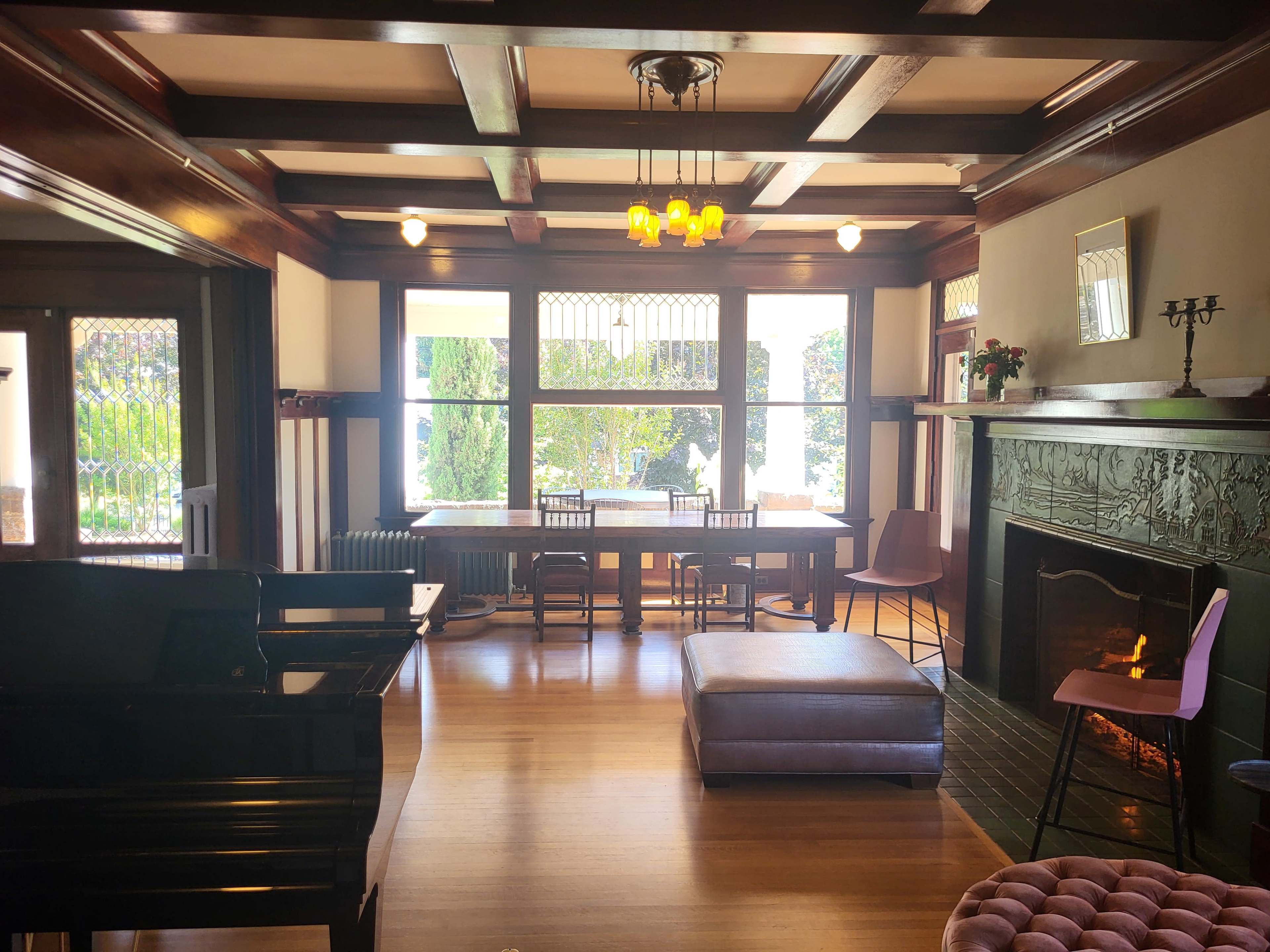 The image shows a spacious living area featuring a black piano, a dining table with chairs, and a fireplace, illuminated by natural light from large windows.