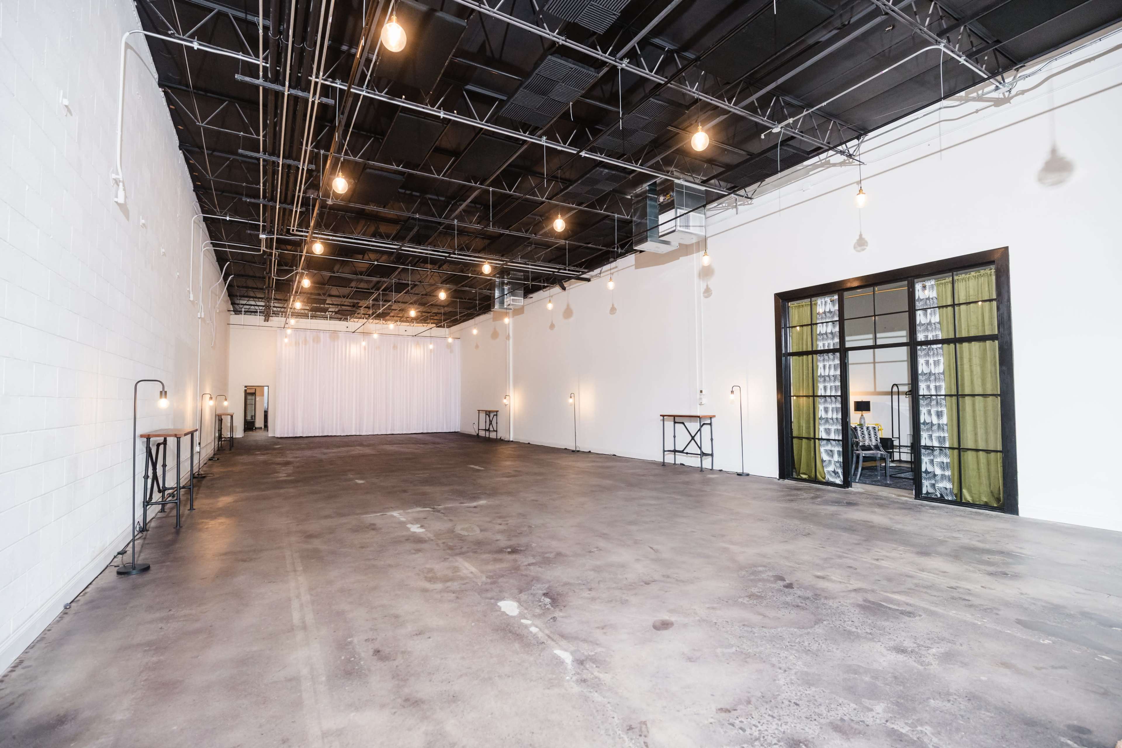 The image shows a spacious, empty industrial-style room with exposed ceilings and warm pendant lighting, featuring a polished concrete floor and white walls.