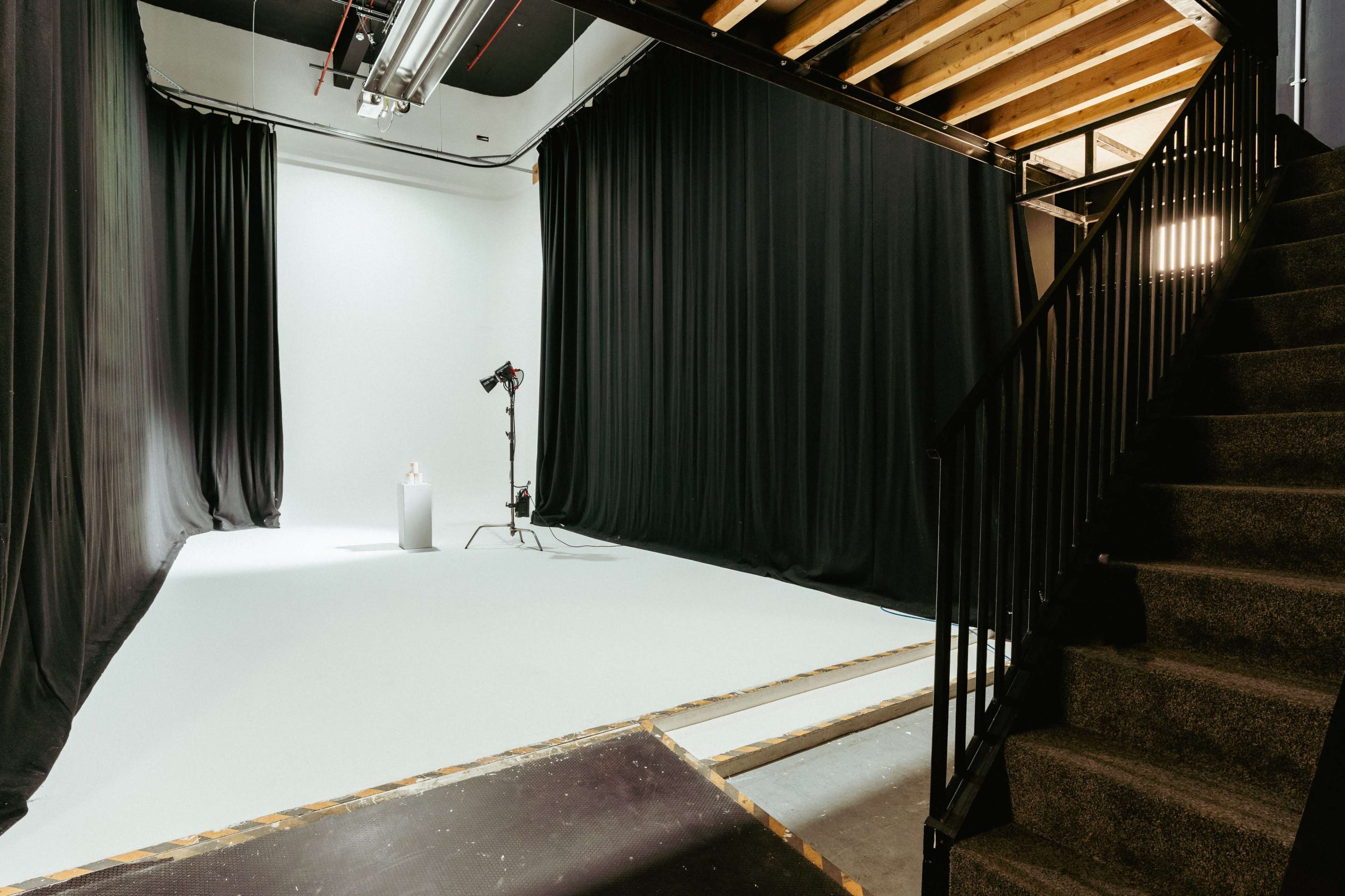 The image shows a spacious photography studio with black curtains, a white backdrop, a light source, and a staircase leading to another level.
