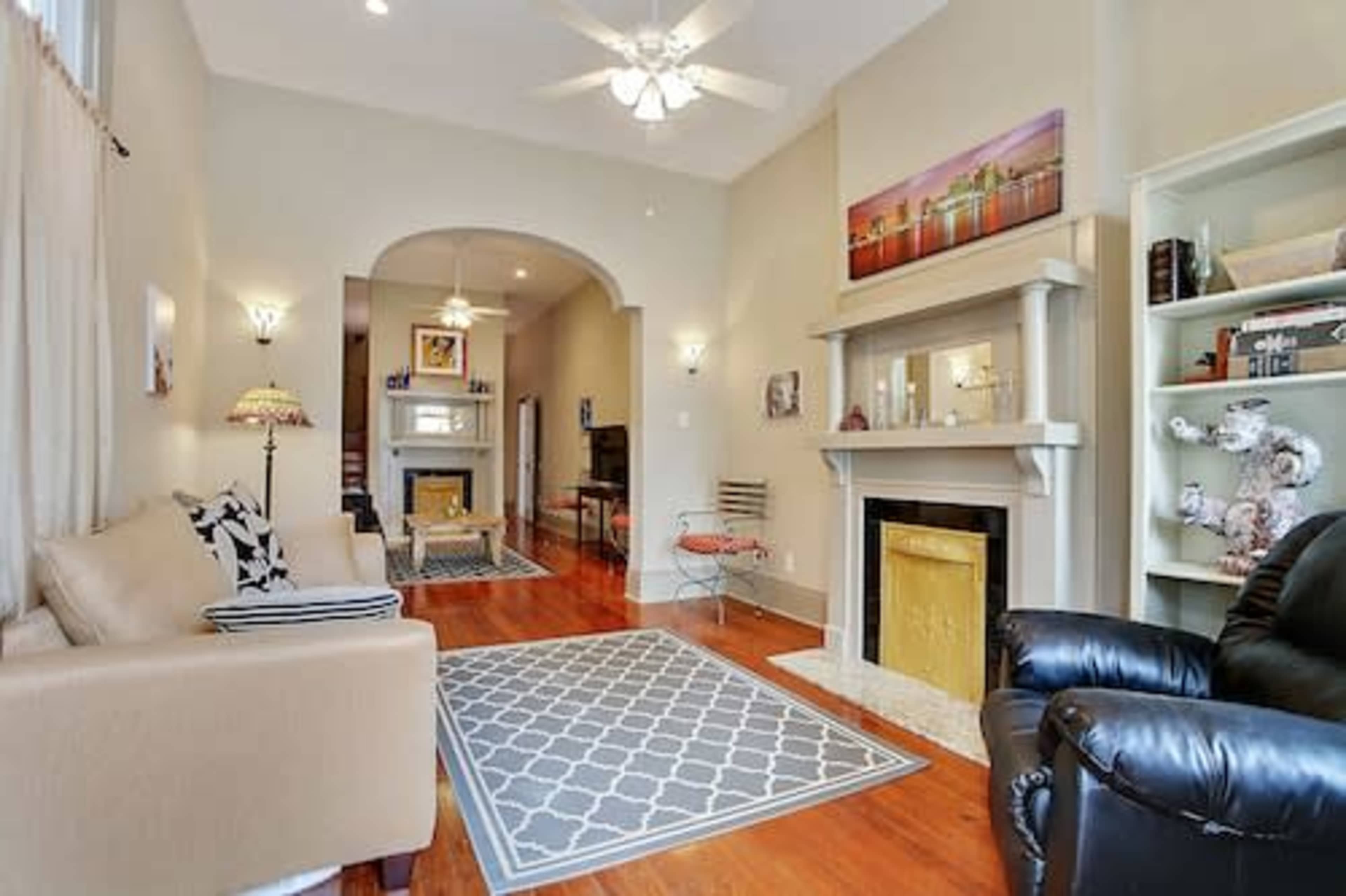 The room features a cozy seating area with a beige sofa and a black recliner, centered around a fireplace with a gold insert, and includes a decorative shelf and a patterned area rug.