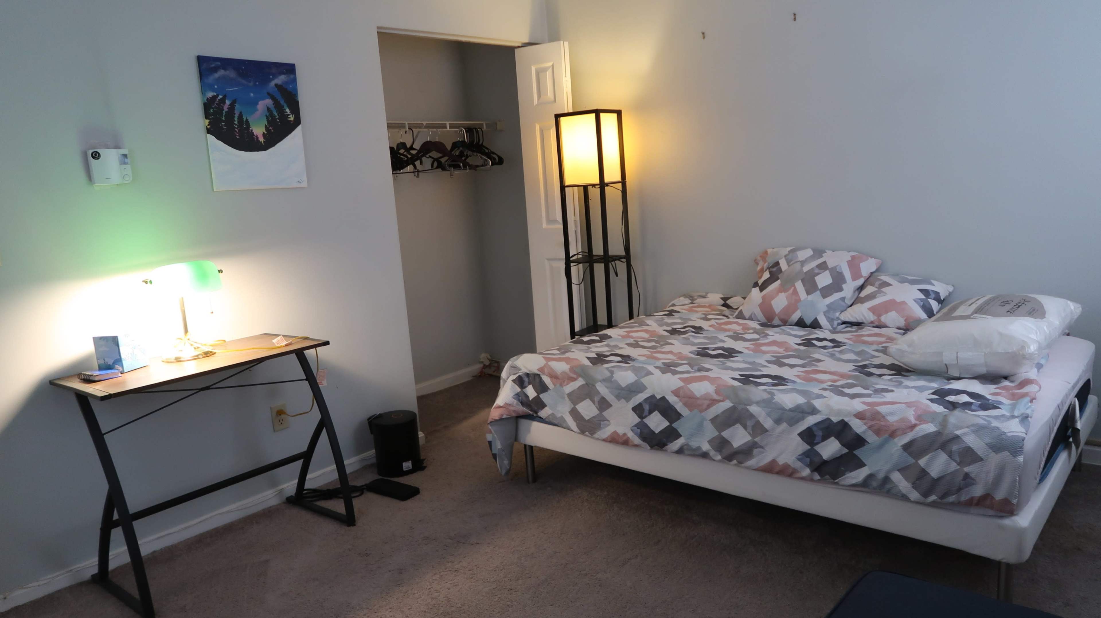 A simple bedroom features a bed with patterned bedding, a desk with a lamp, and a closet in the corner.