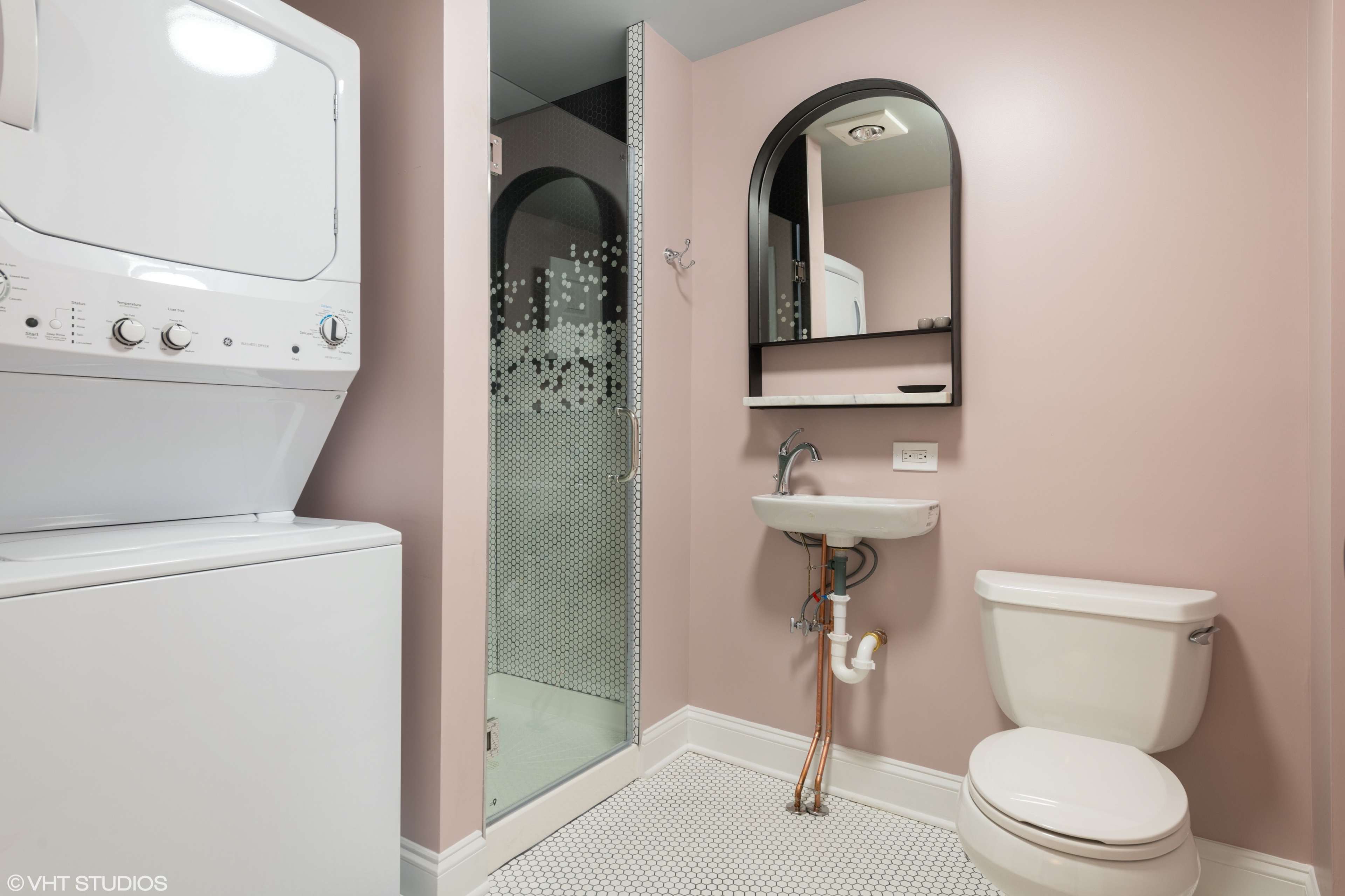 A compact laundry room features a stacked washer and dryer, a small sink with a wall-mounted mirror, and a shower stall, all set against pink walls.