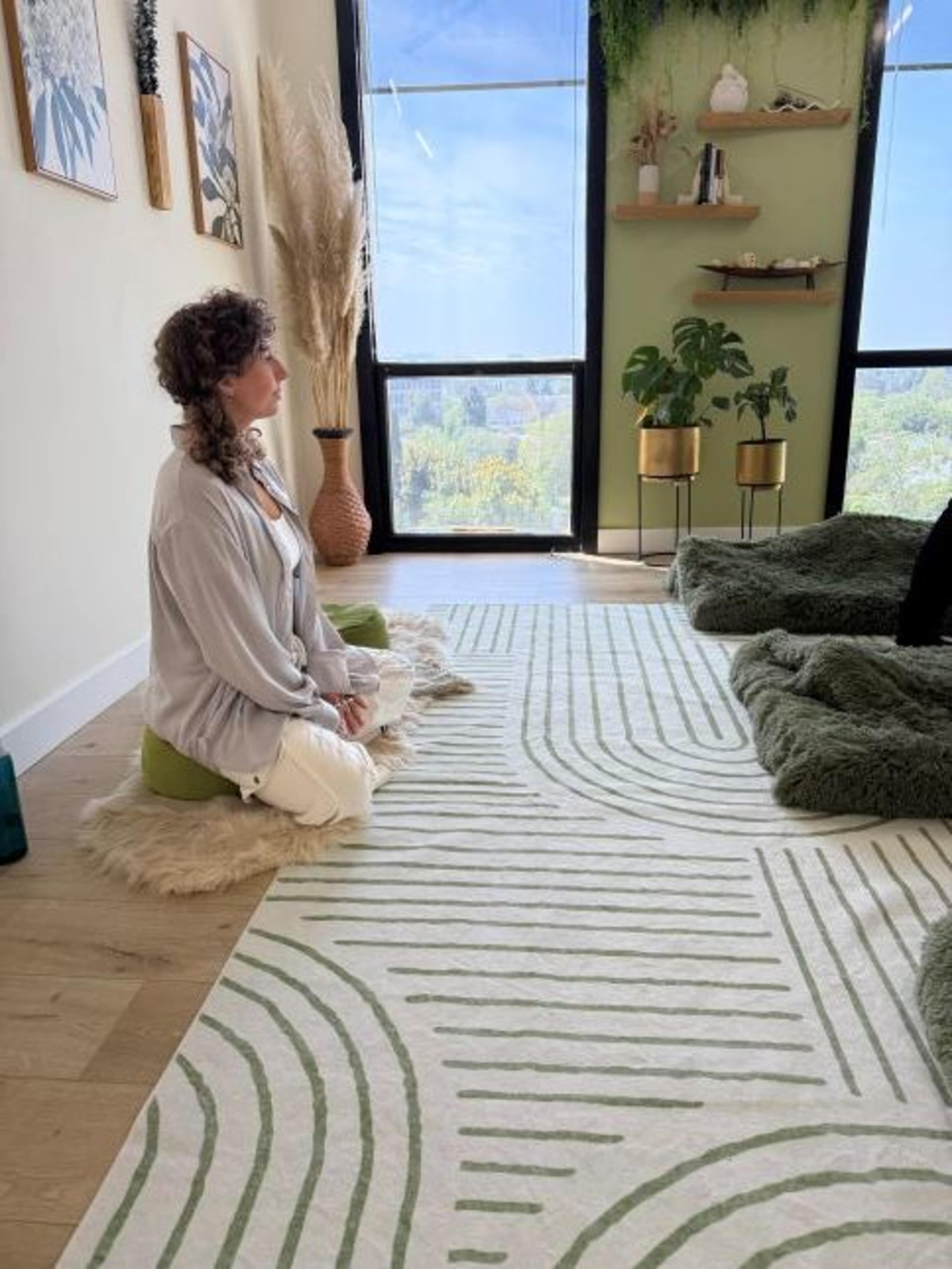 A person sits cross-legged on a rug in a serene room with large windows, soft greenery, and minimalist decor.