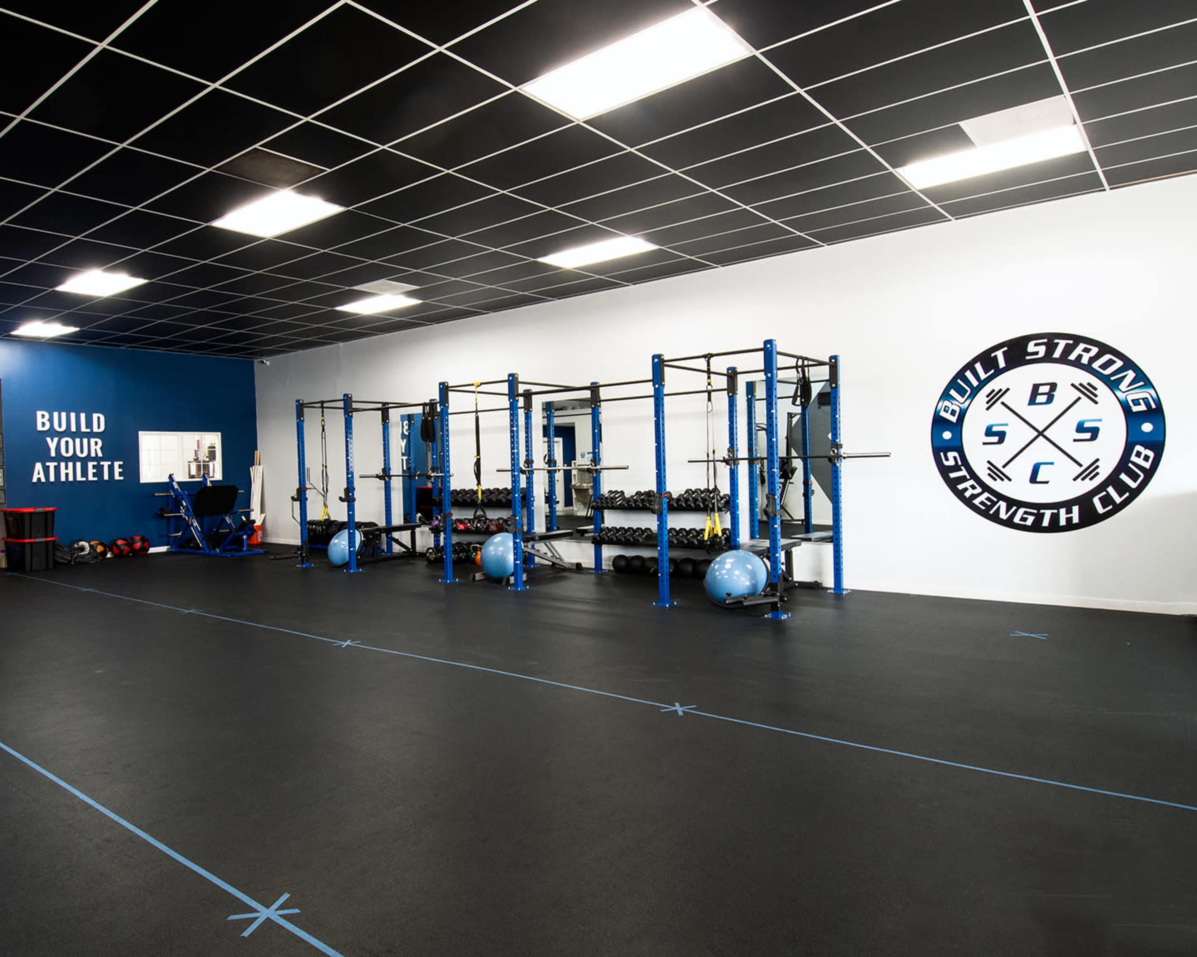 A well-equipped gym featuring weightlifting racks, dumbbells, and exercise balls, with the words "BUILD YOUR ATHLETE" on one wall.