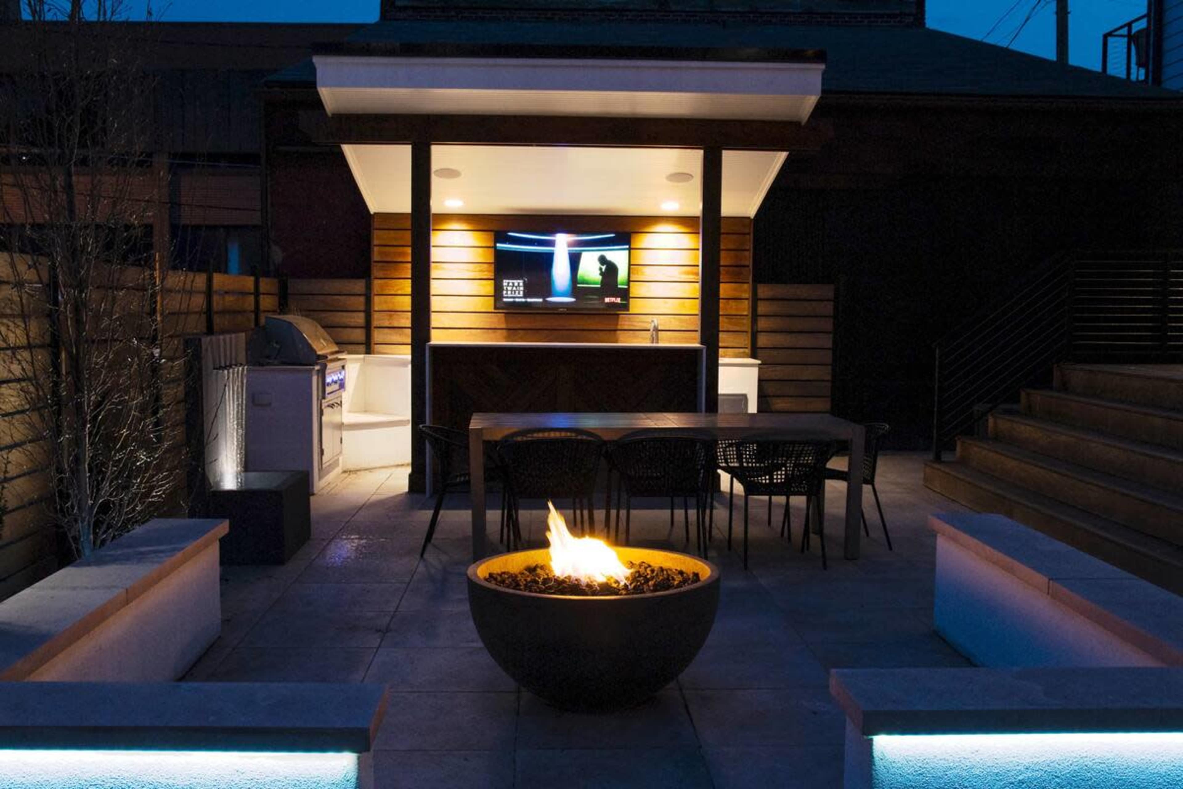 An outdoor patio area at night featuring a fire pit, a dining table, an outdoor kitchen with a grill, and a television mounted on a wooden structure.