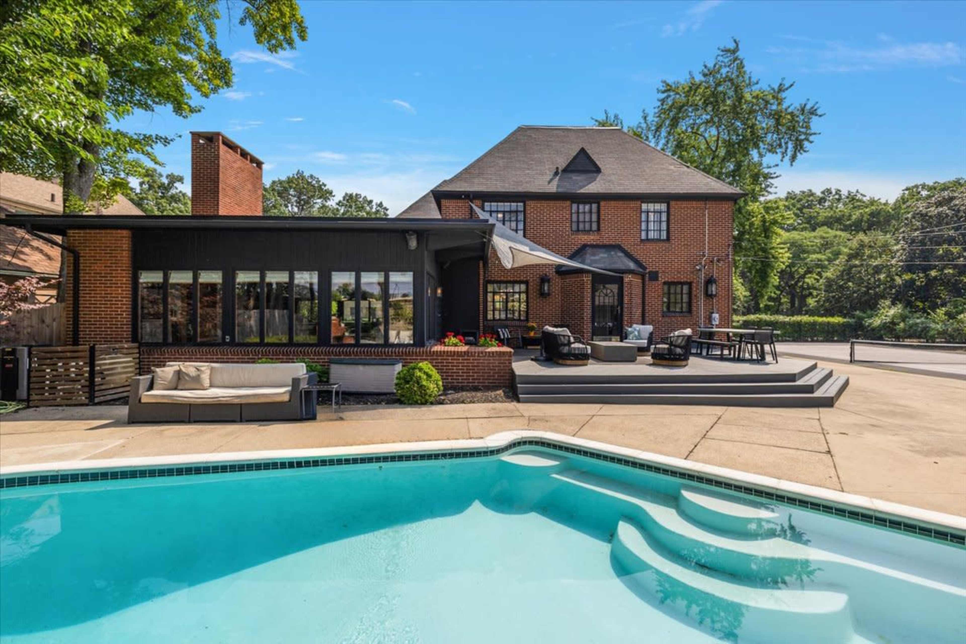 The image shows a backyard with a swimming pool in the foreground and a two-story brick house with a landscaped area in the background.