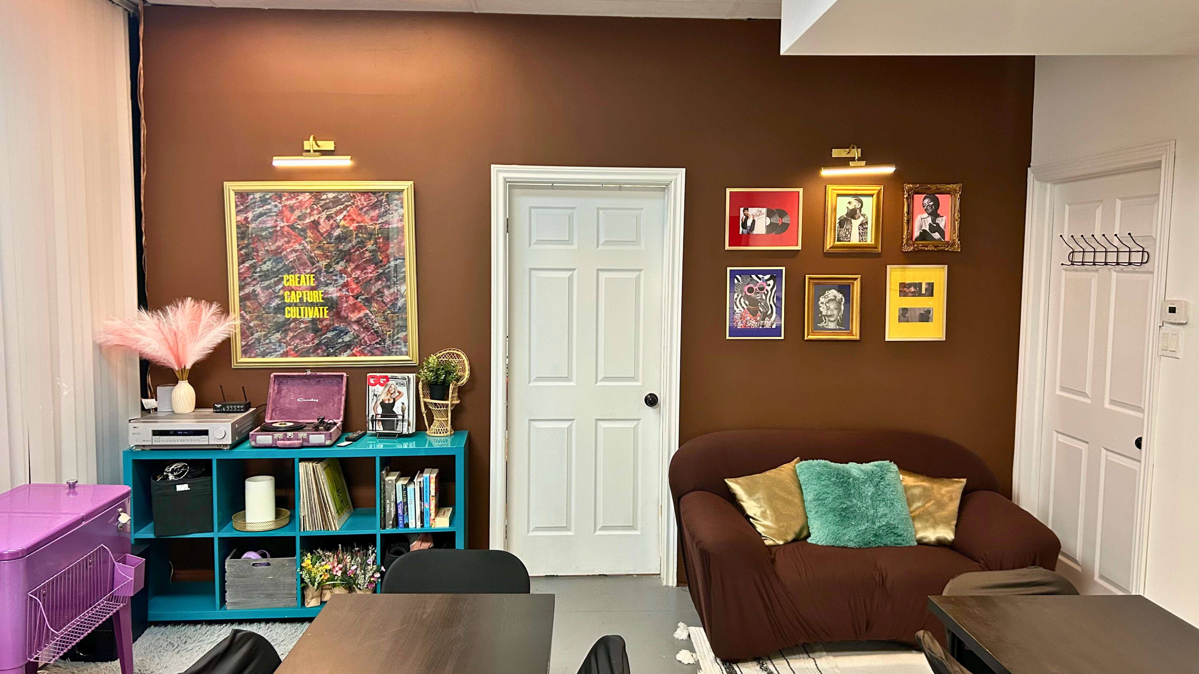The image shows a cozy interior with a brown accent wall, featuring framed artwork and a teal bookshelf, alongside a brown sofa and decorative plants.