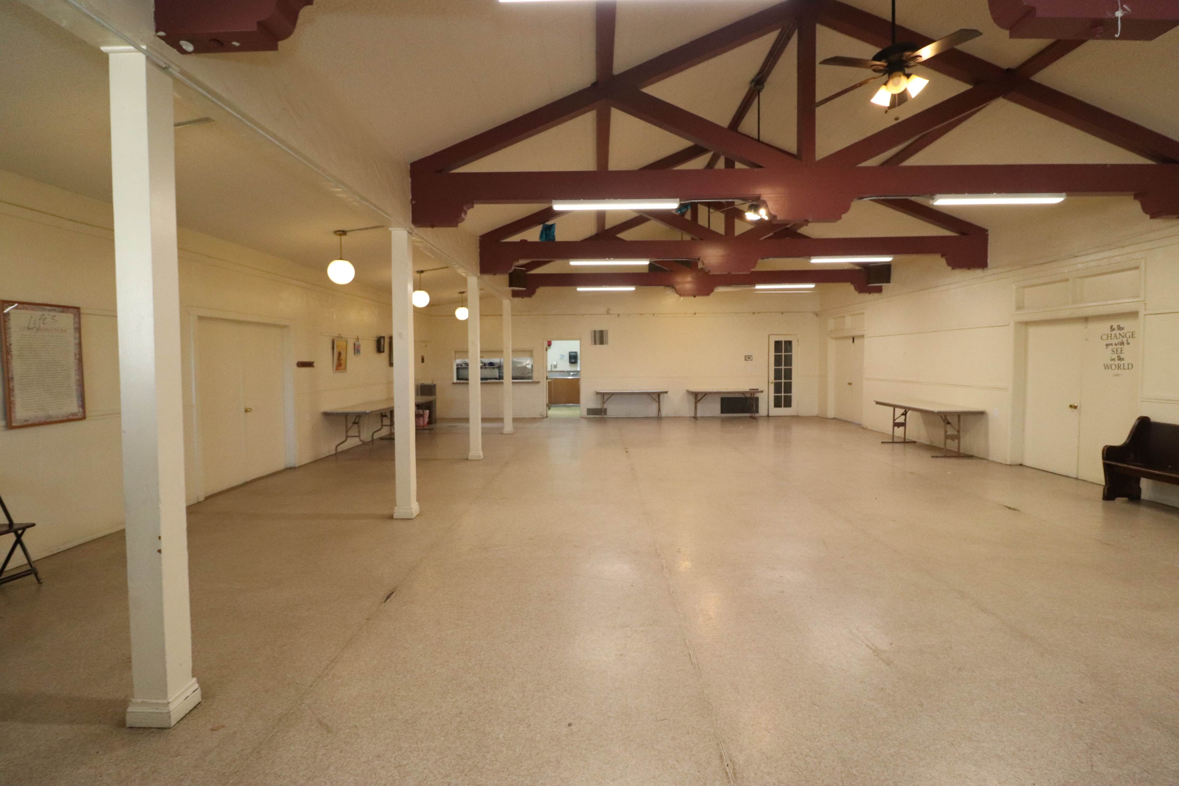 A large, empty room with a high ceiling, exposed wooden beams, and several tables along the walls.
