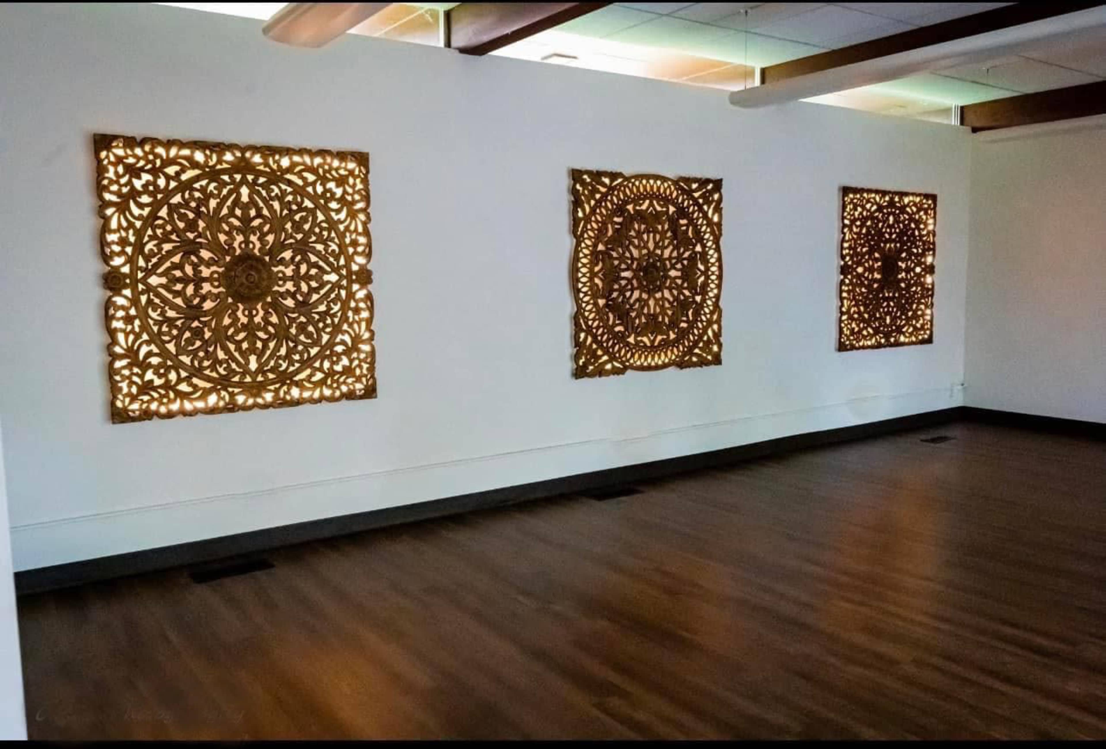 Three intricately designed wooden panels illuminated from behind, mounted on a white wall in a spacious room with wooden flooring.