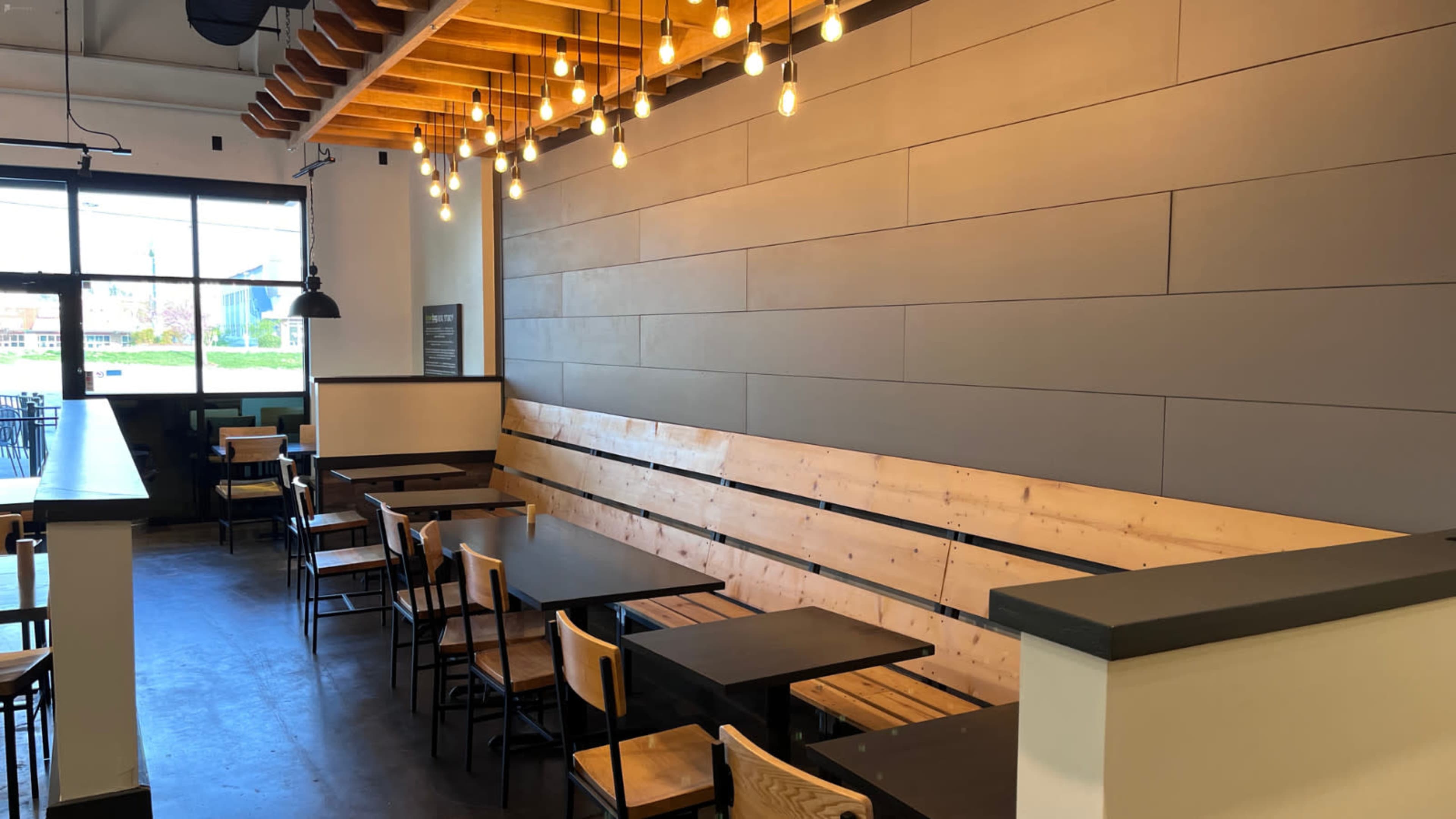 An interior of a restaurant featuring wooden benches along a wall, black tables, and hanging light fixtures.