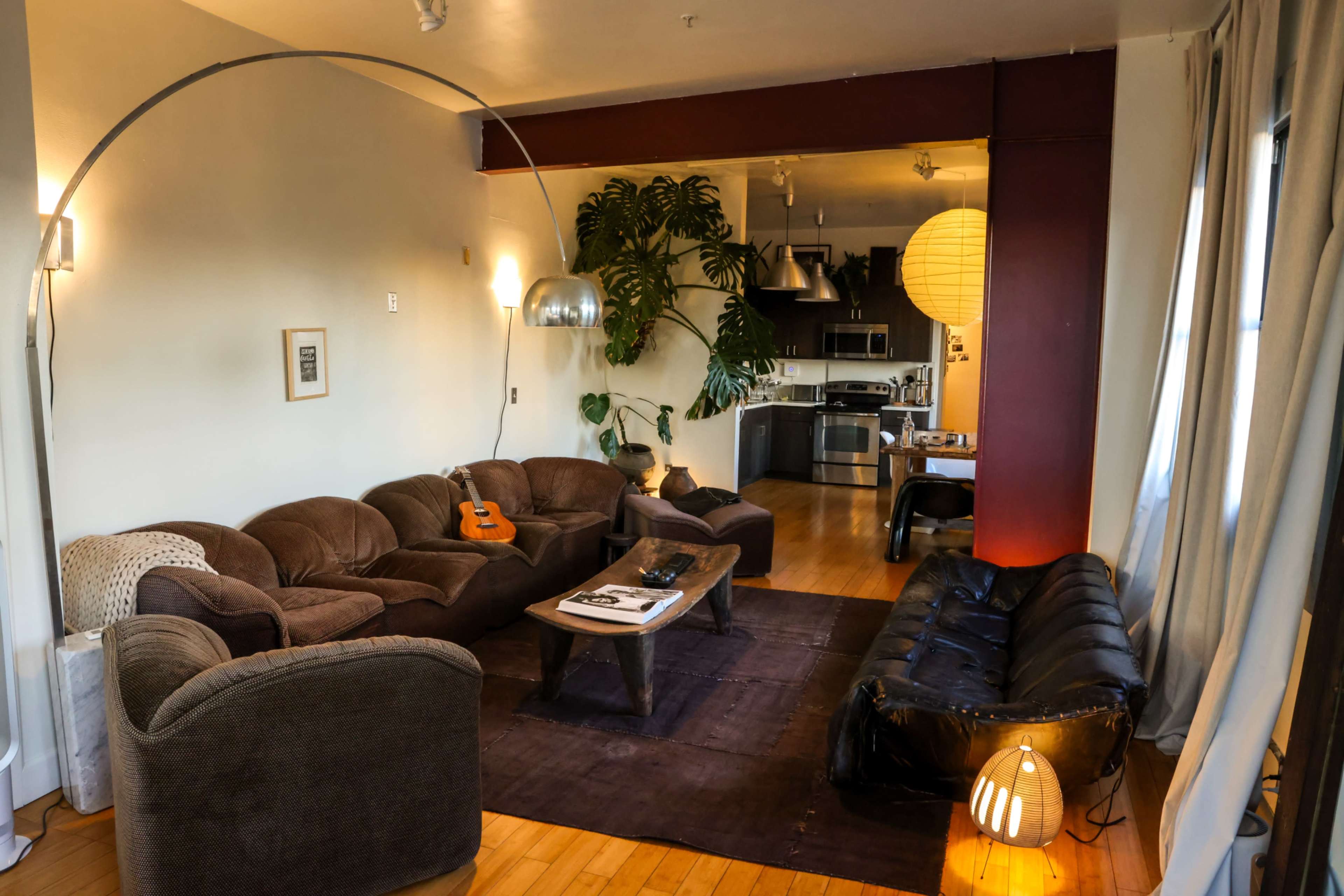 The image shows a contemporary living room with several seating areas, a wooden coffee table, and a kitchen visible in the background.