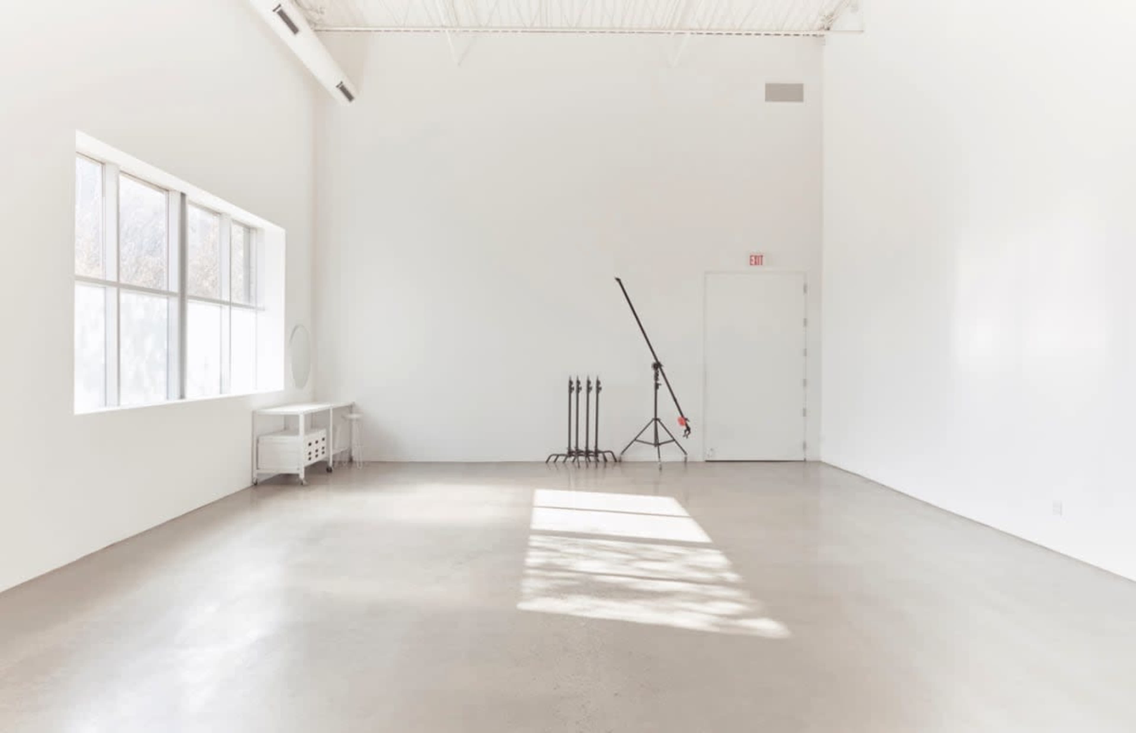 The image shows a minimalistic, empty room with white walls and a concrete floor, featuring large windows and a standing lamp in the corner.