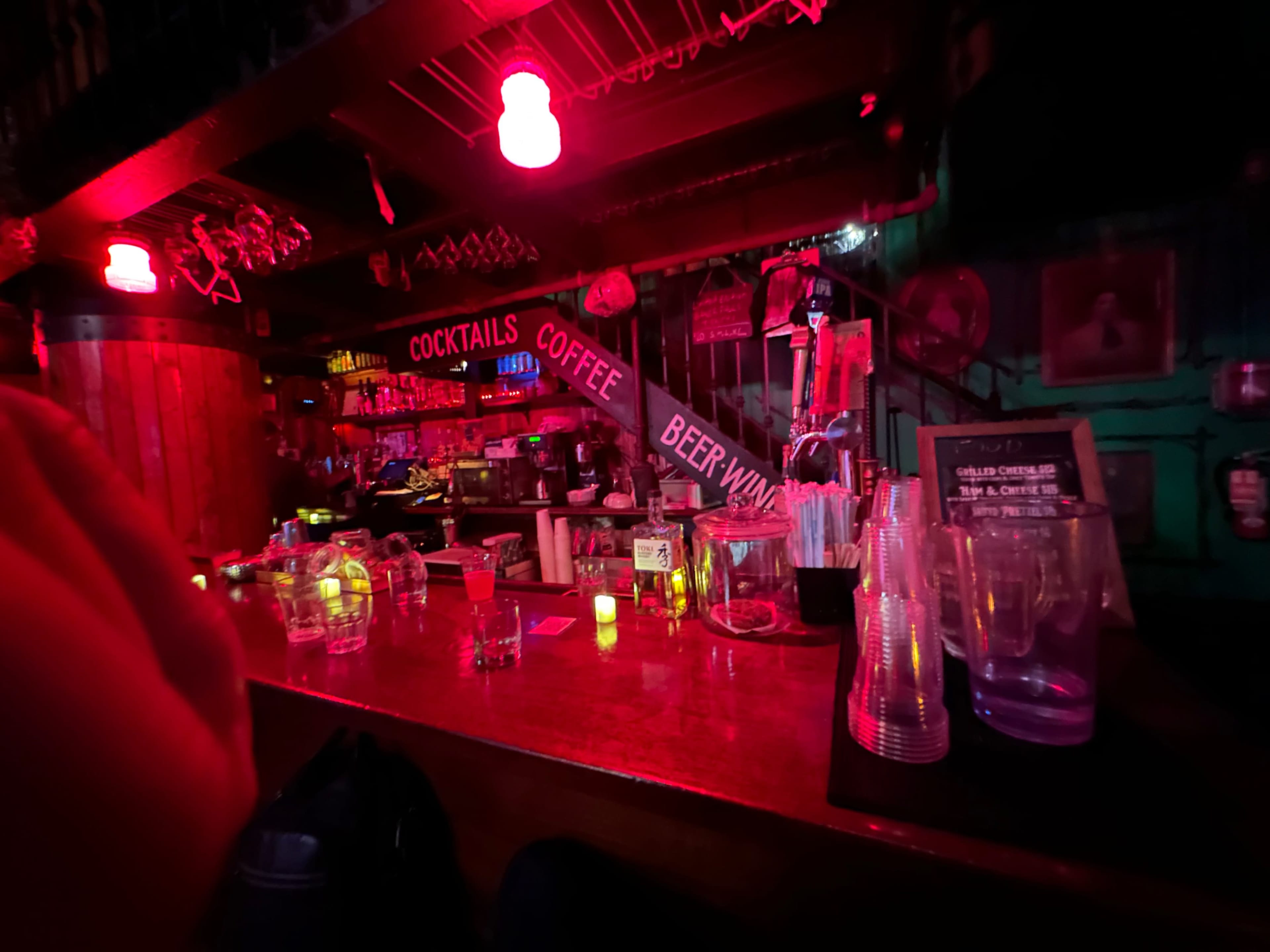 The image shows a dimly lit bar with red lighting, featuring various bottles and glasses on the shelves behind the counter.