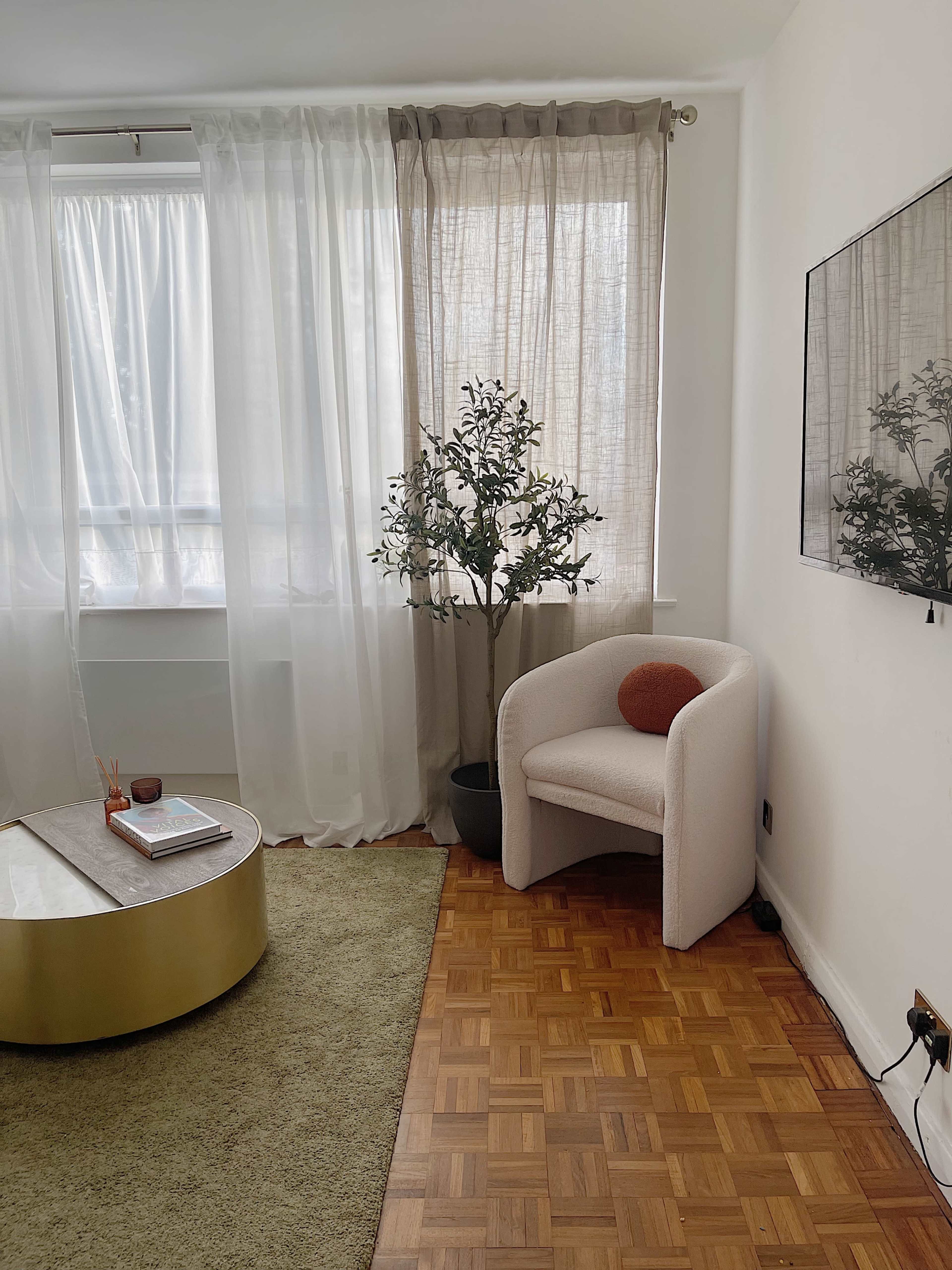 A cozy living room with a light-colored chair, a round coffee table, a decorative plant, and large sheer curtains.