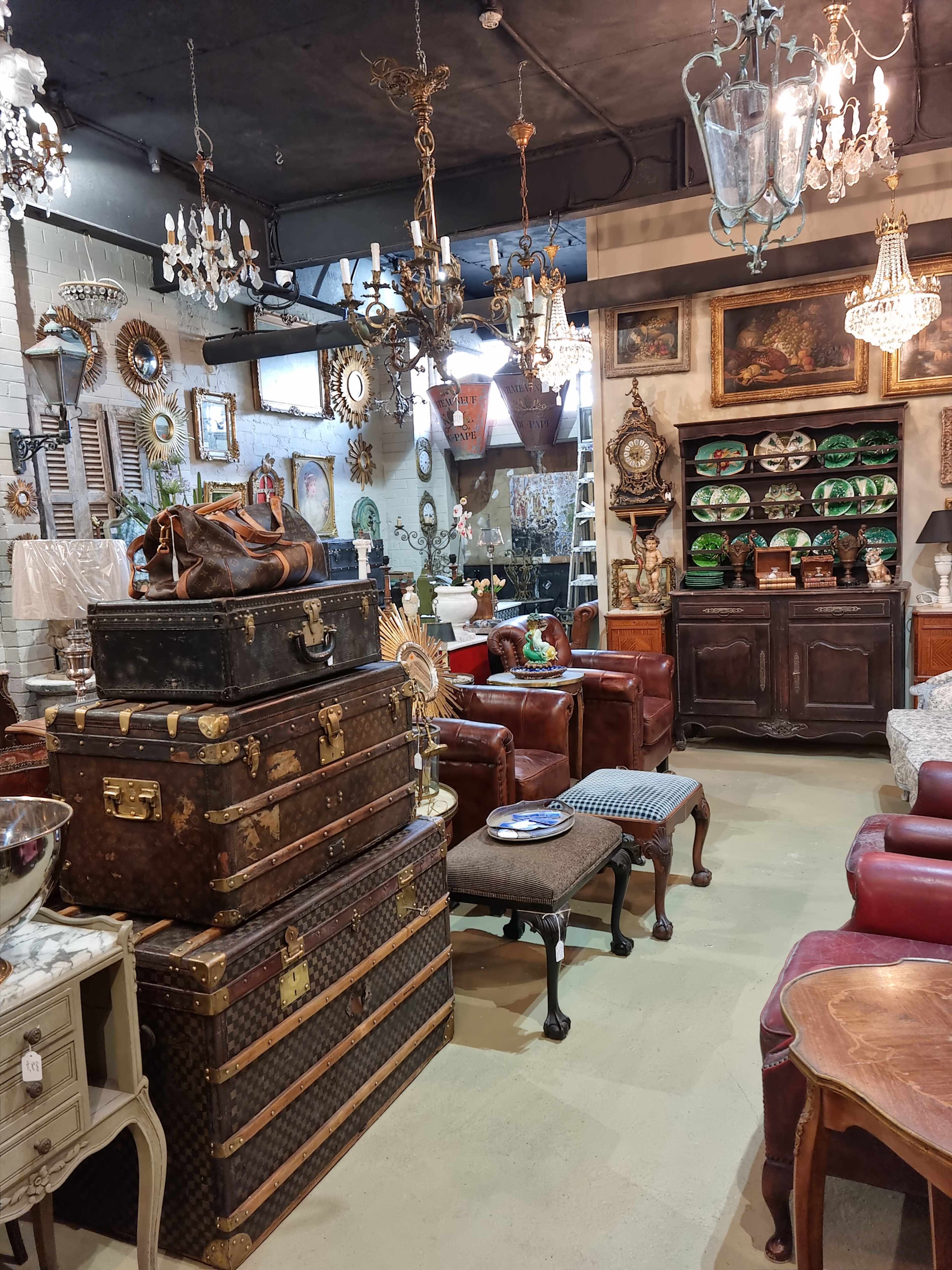 A vintage shop interior filled with various antique furniture, decorative items, and chandeliers, arranged around leather sofas and stacked trunks.