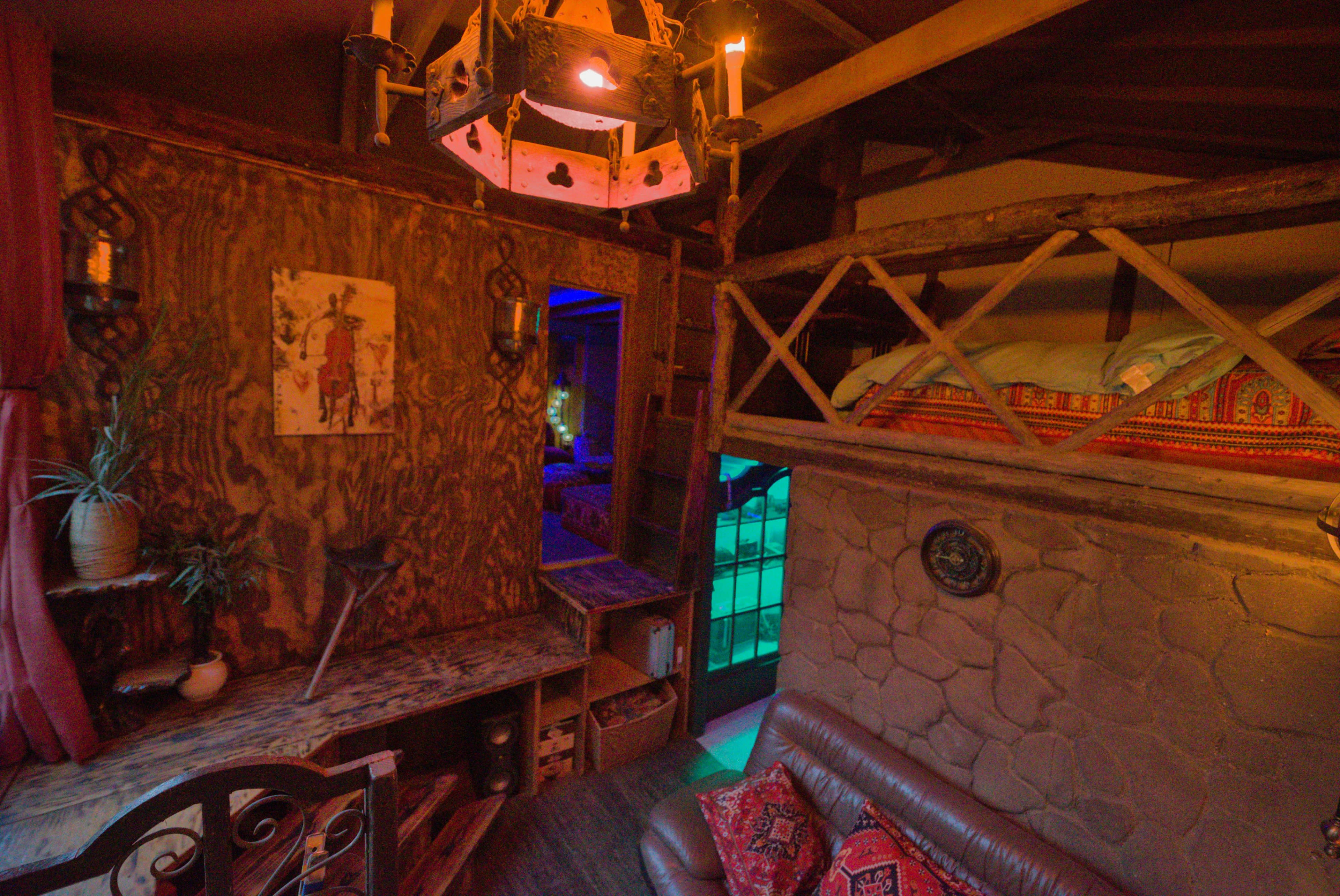 The image shows a cozy interior of a room with a wooden loft, a stone wall, and various decorative elements, including a chandelier and a sofa with patterned cushions.