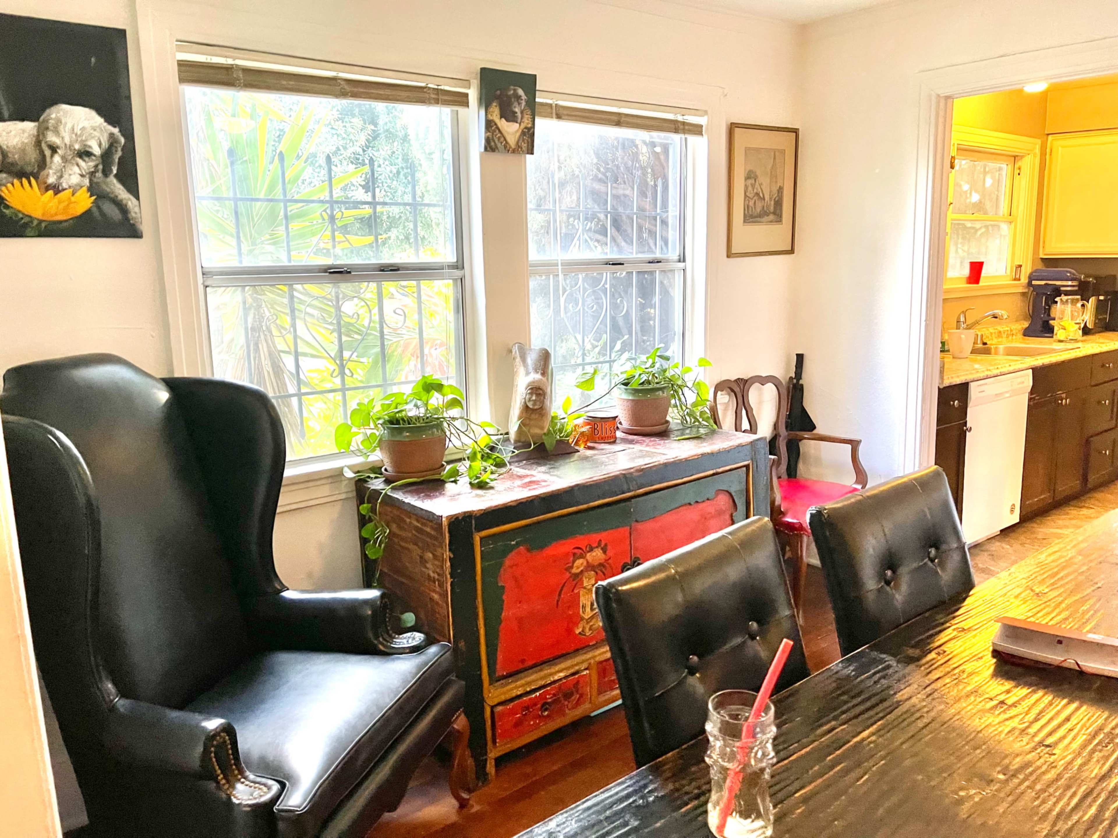 A dining area features a large dark wooden table, a black armchair, a colorful vintage chest, and plants by the windows.