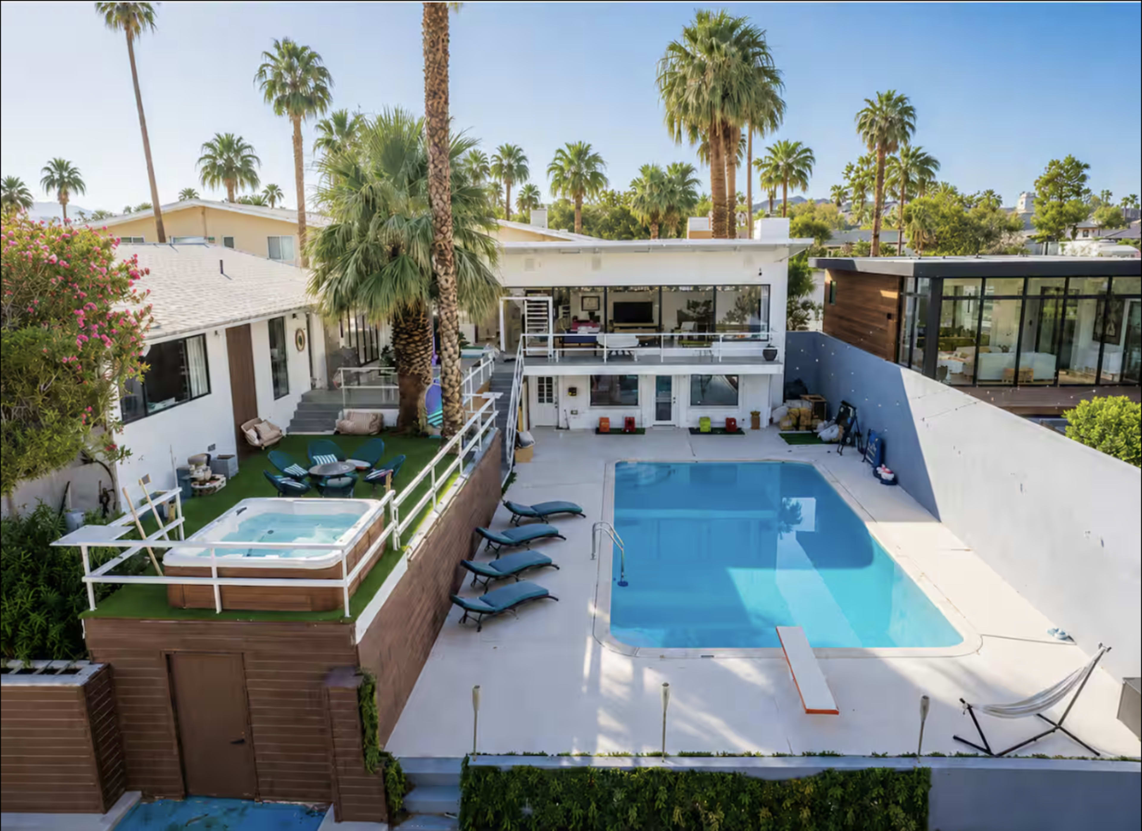 The image shows a modern backyard with a large pool, lounge chairs, and a hot tub surrounded by palm trees and a contemporary house.