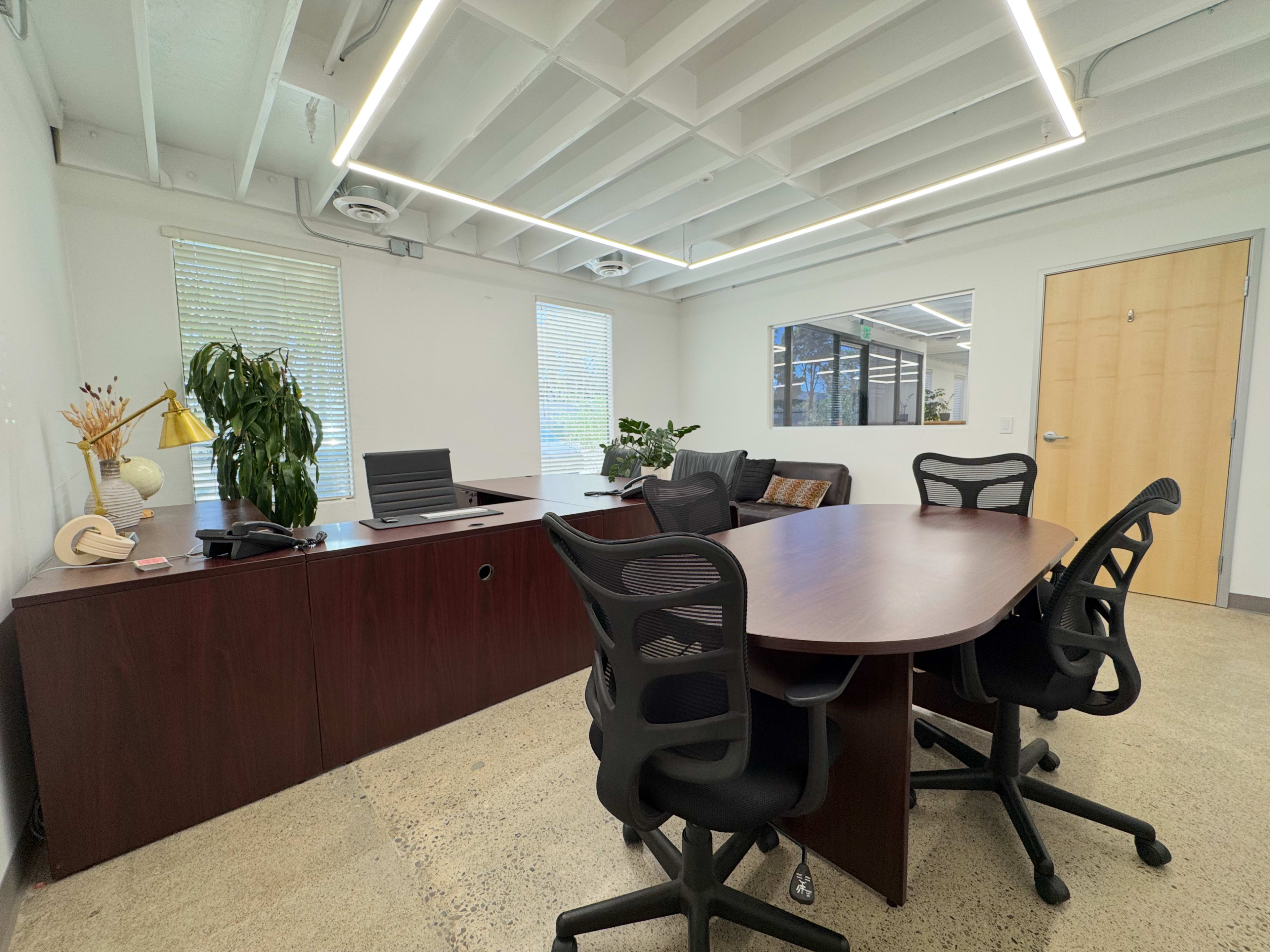The image shows a modern office space featuring a large wooden desk, a conference table with four black chairs, and seating along with potted plants.