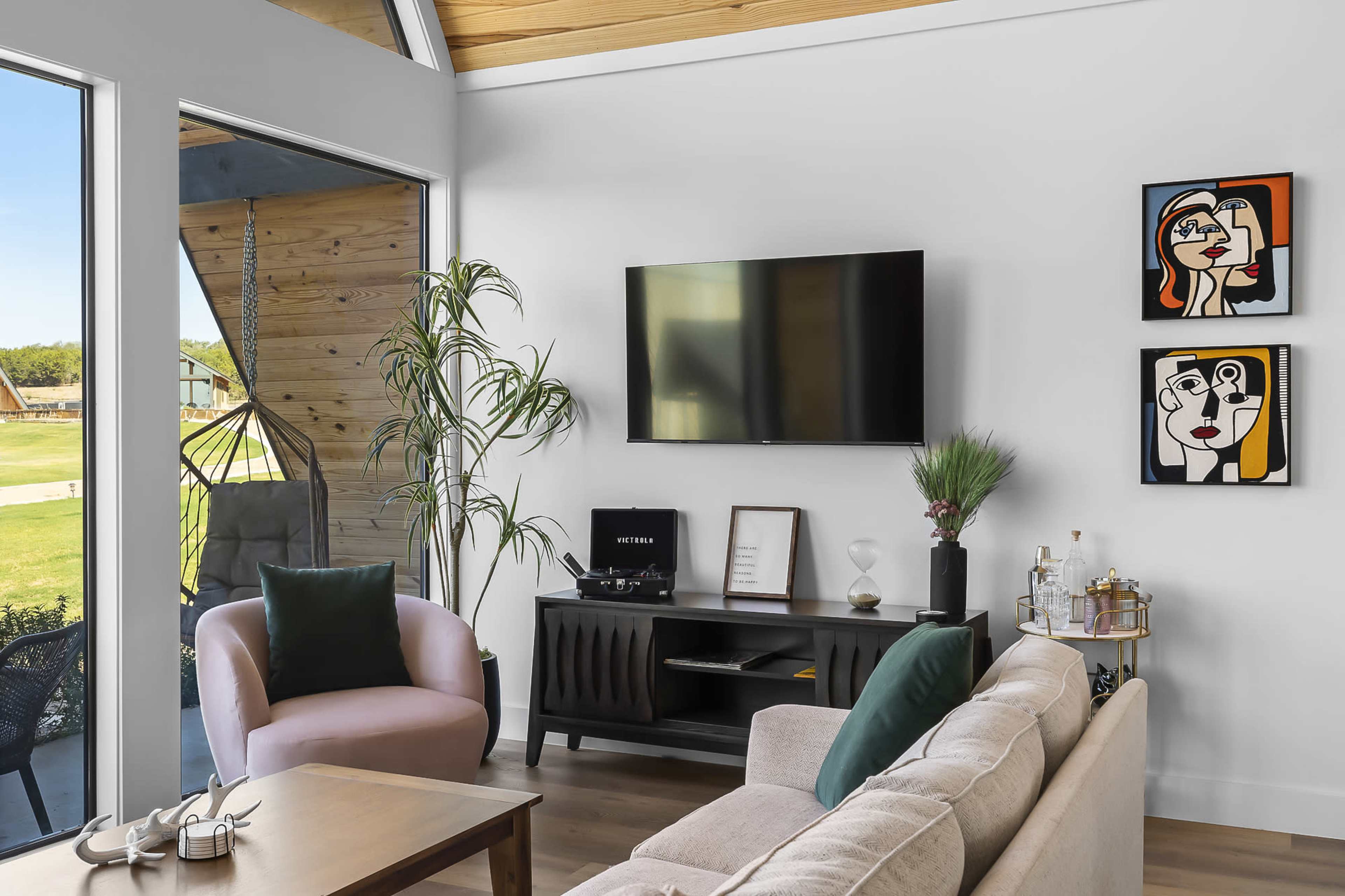 A modern living room features a light-colored sofa, a black television mounted on the wall, and two framed artworks beside a window with a view of the outdoors.