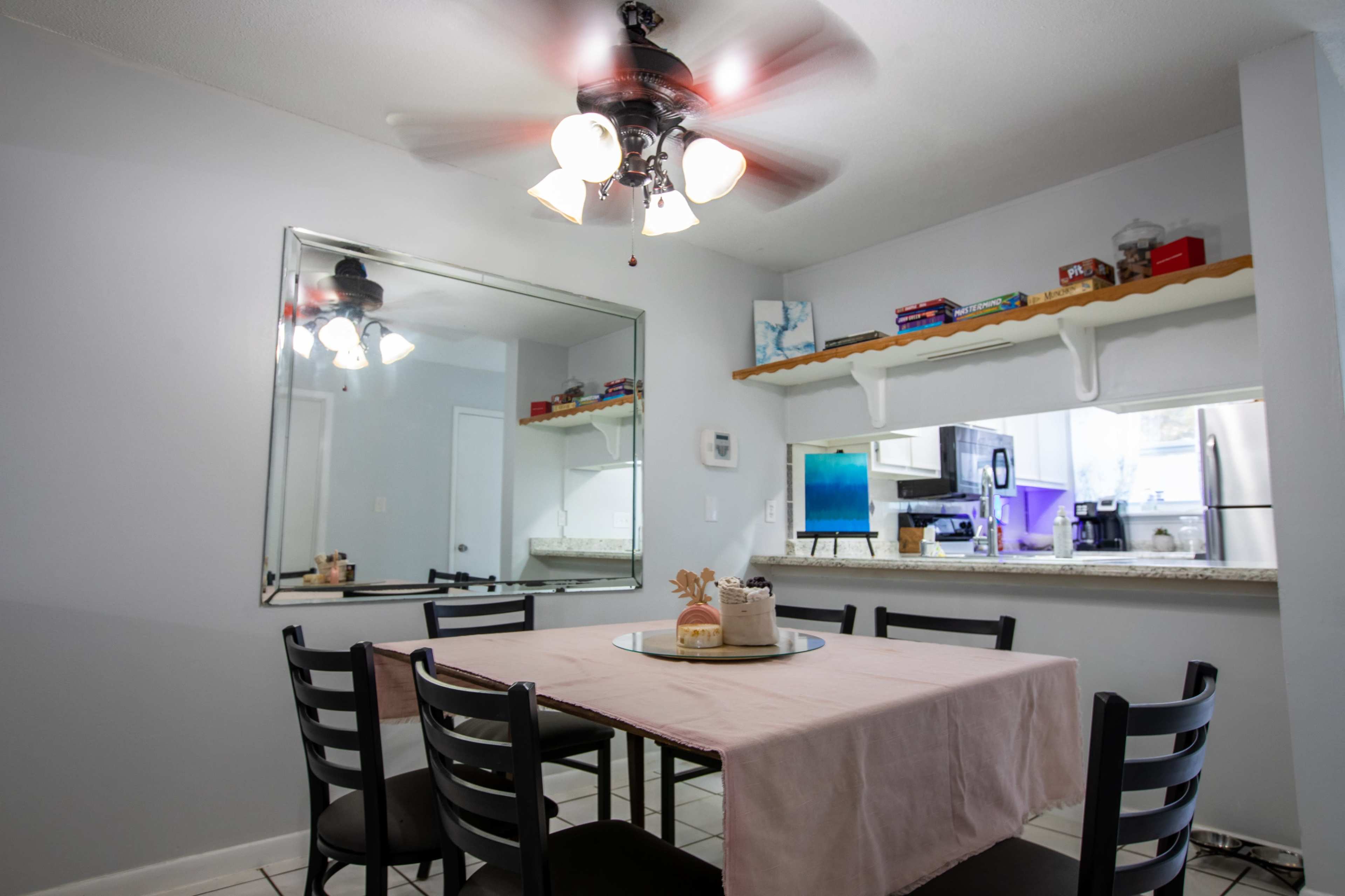 A dining area features a table set with a neutral tablecloth, surrounded by black chairs, with a ceiling fan above and a large mirror on the wall reflecting a kitchen space.