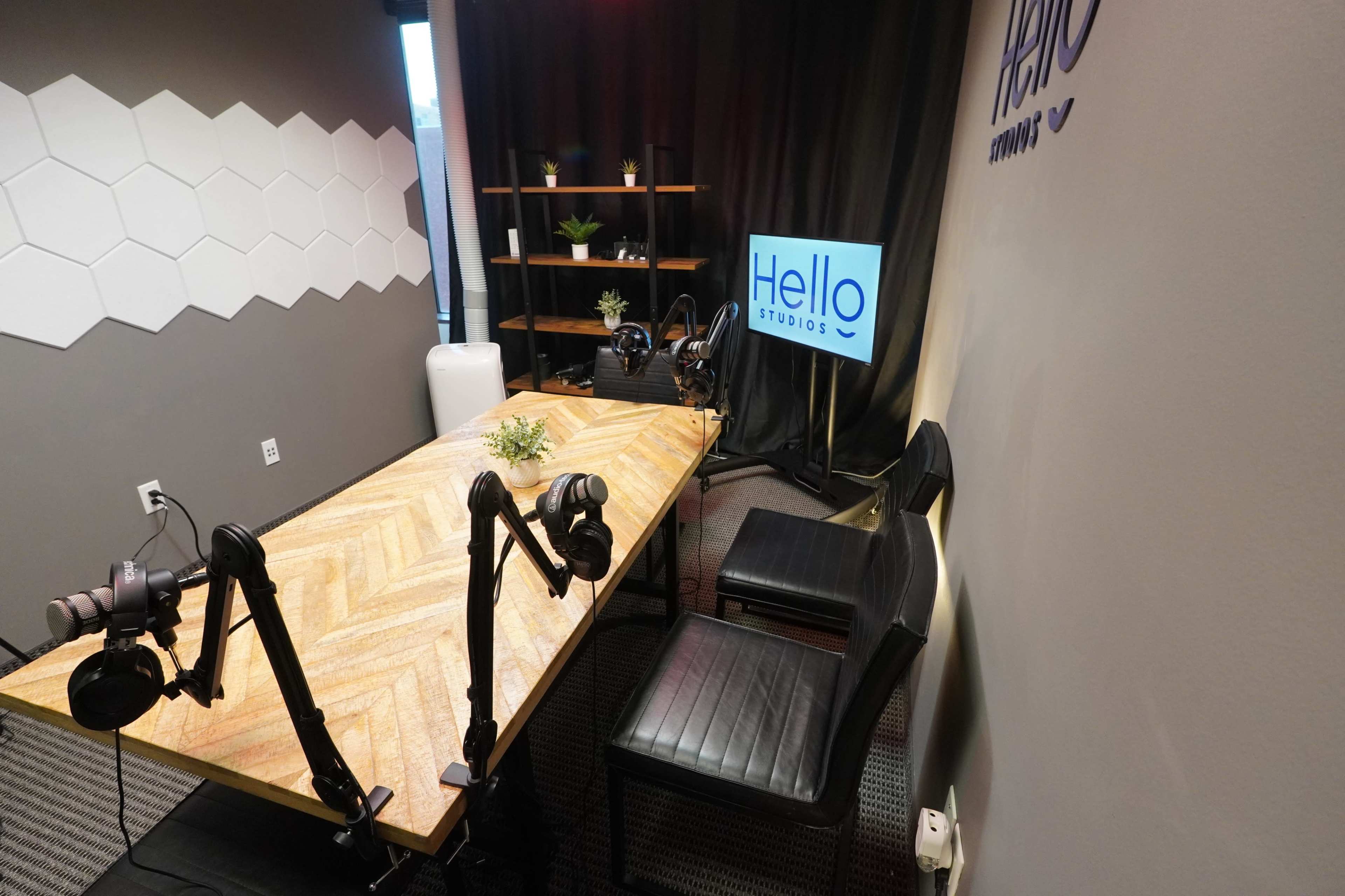 The image shows a podcast recording studio with a wooden table, two black chairs, microphones on stands, and a television display with the "Hello Studios" logo.