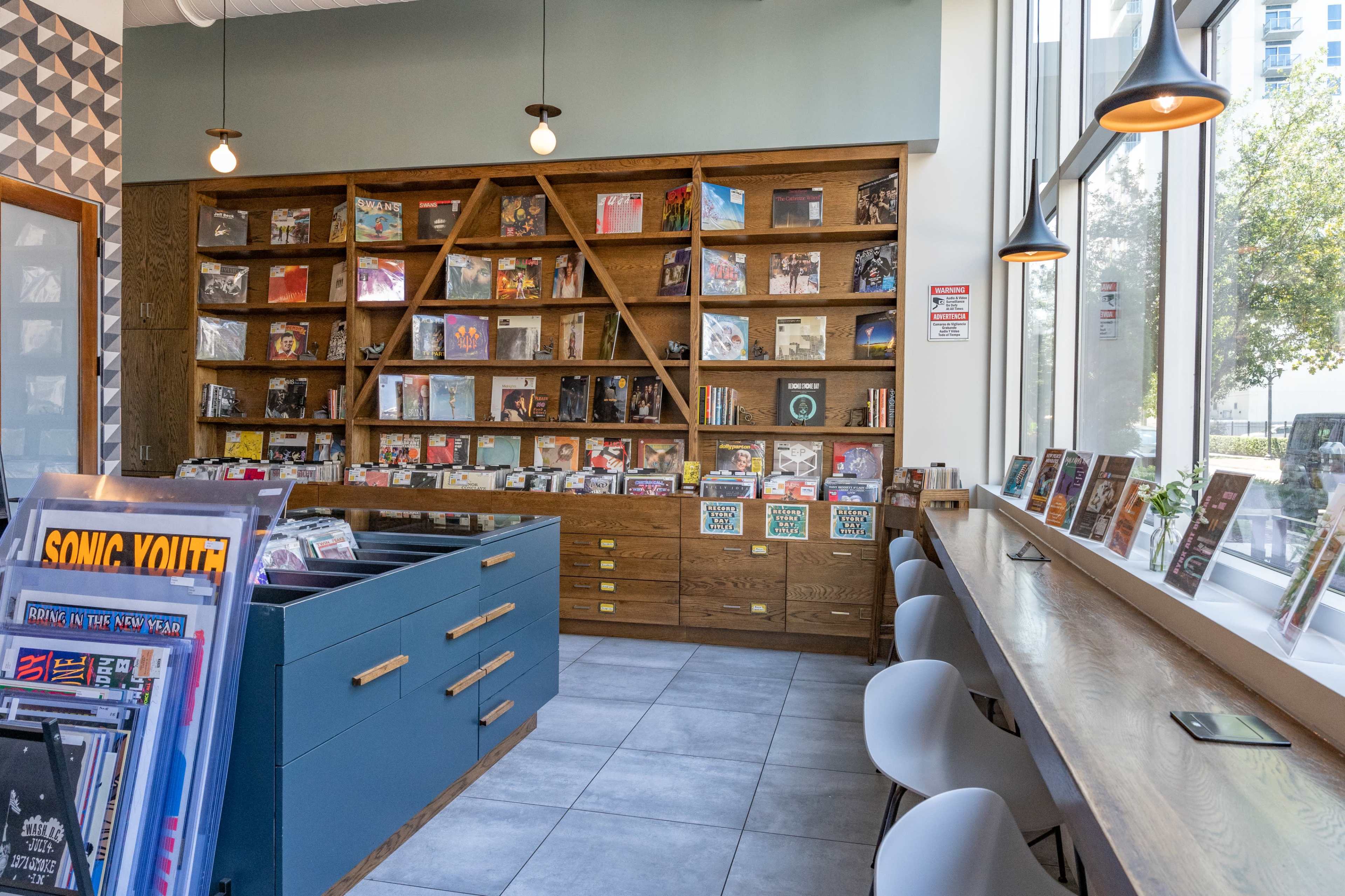 The image shows a modern space with wooden shelves filled with vinyl records, a blue display counter, and a seating area with white chairs.