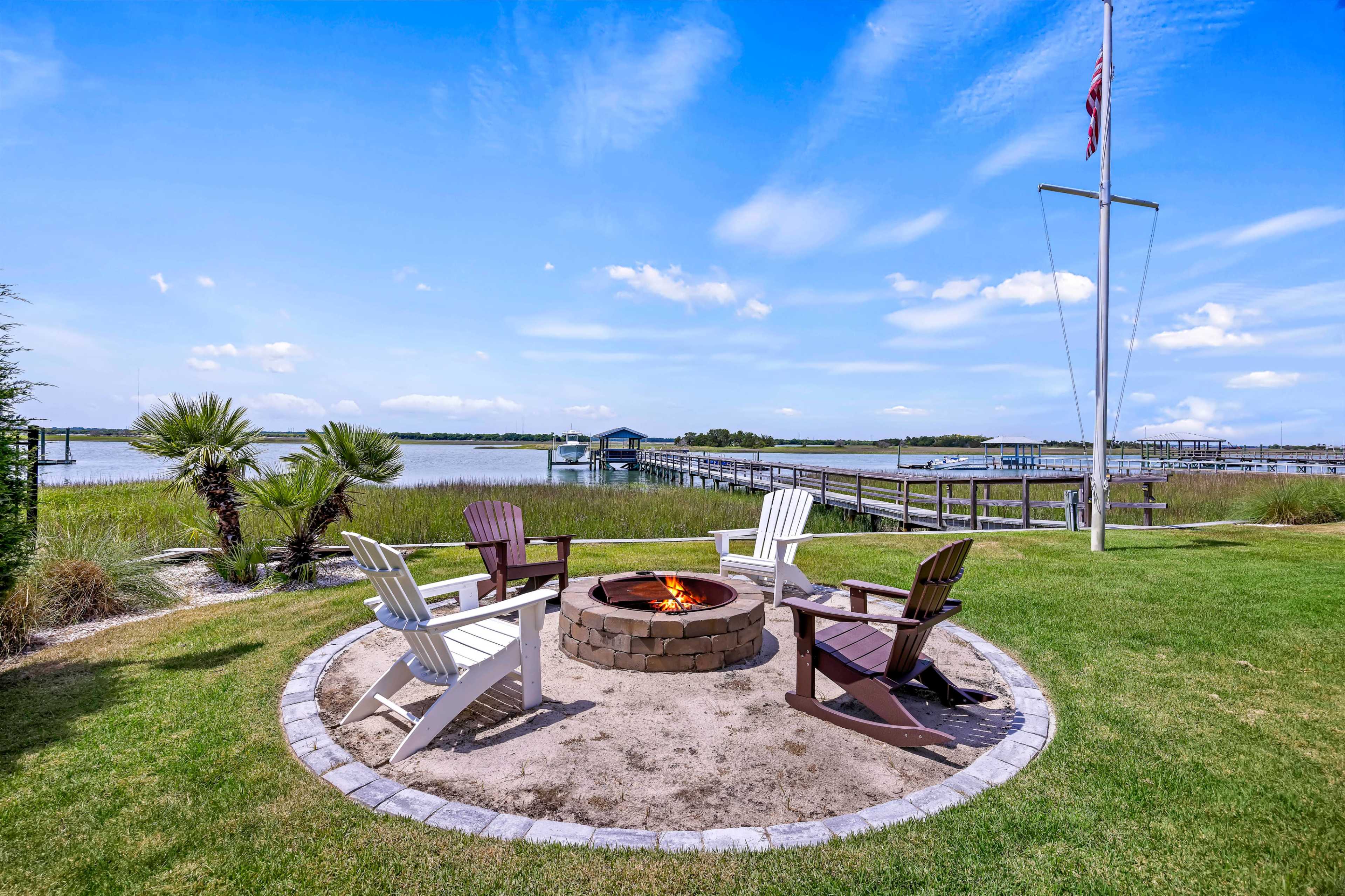 A circular fire pit surrounded by four Adirondack chairs is situated near a lake, with a wooden dock visible in the background.