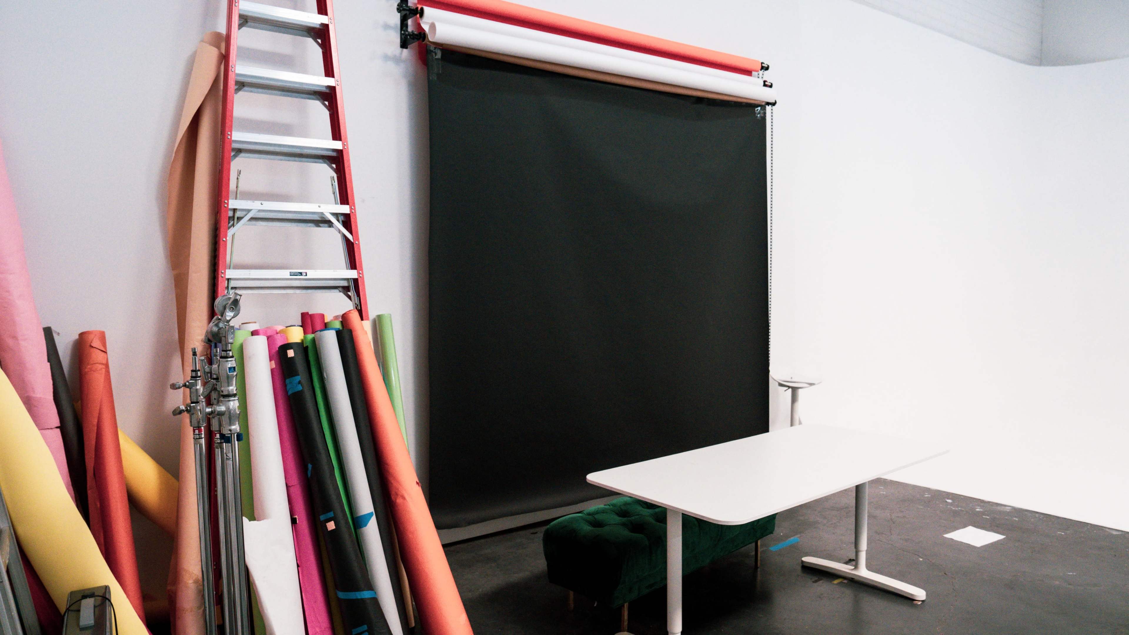 A studio space features a white wall, a table, a green upholstered bench, a multicolored backdrop roll, and a ladder leaning against the wall.