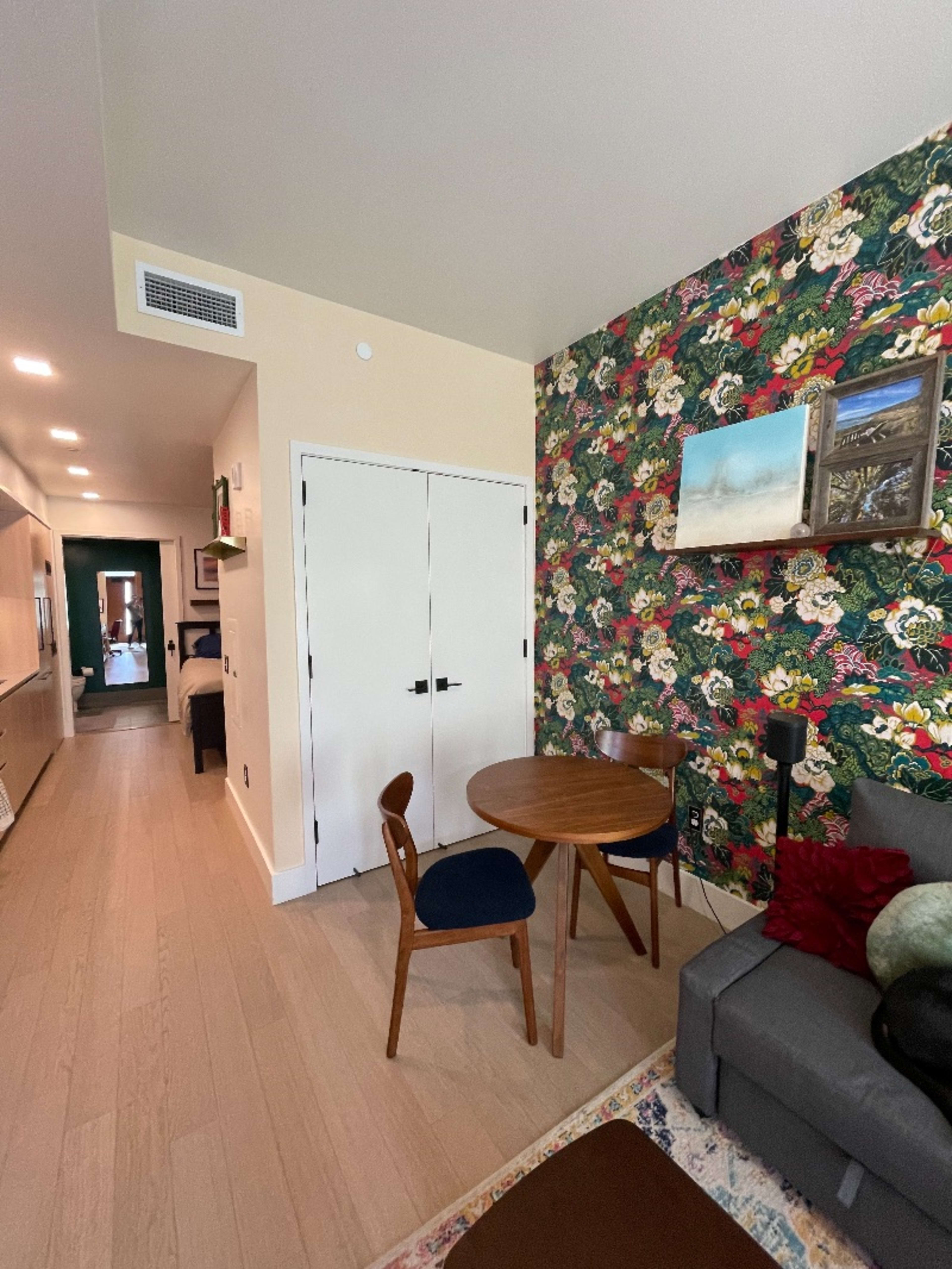 A compact living space featuring a round wooden table with a blue chair, a gray sofa, and a floral wallpaper accent wall.