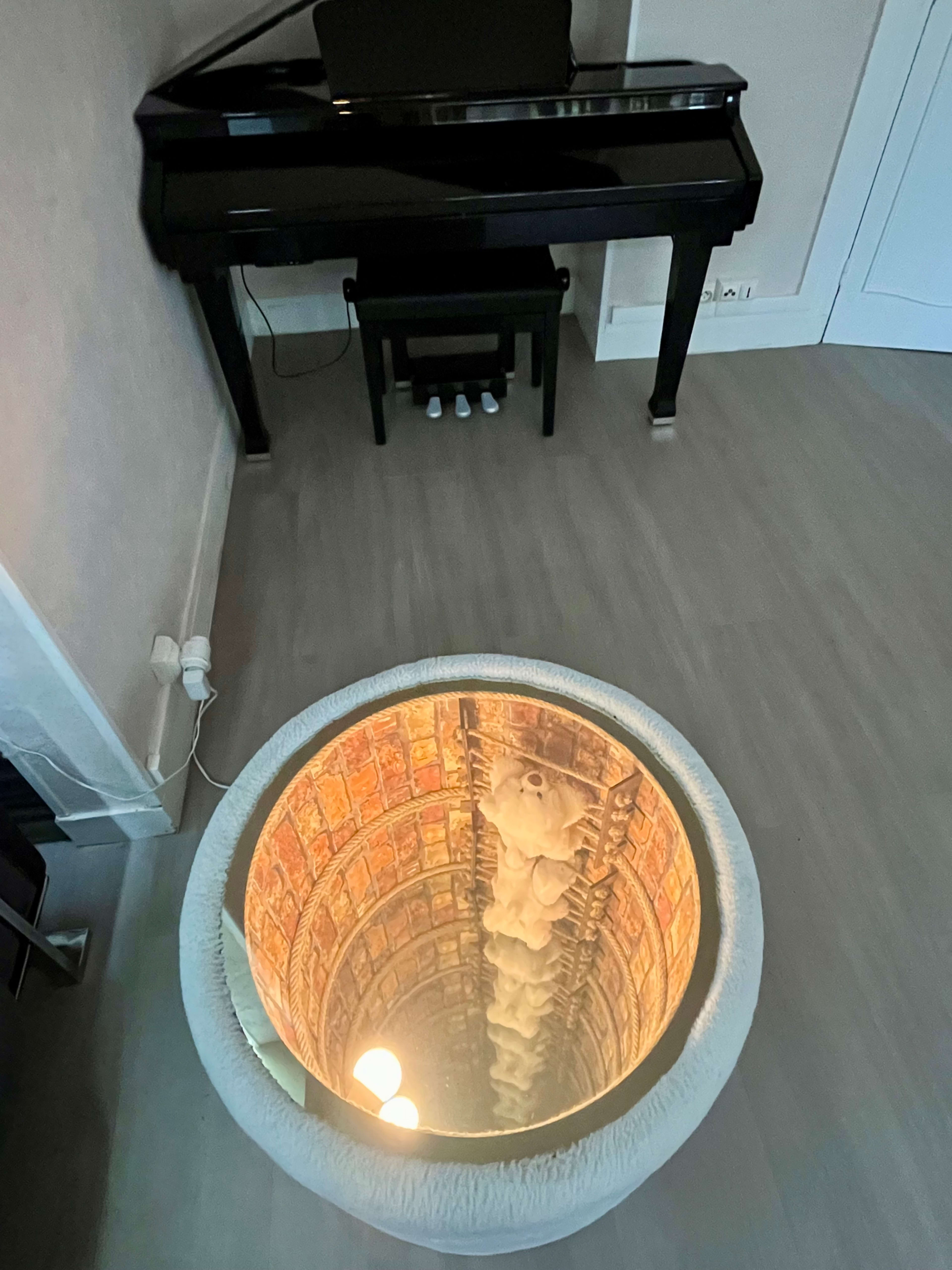 A black grand piano is positioned next to a circular hole in the floor, revealing an intricate, downward view of a spiral stairwell.