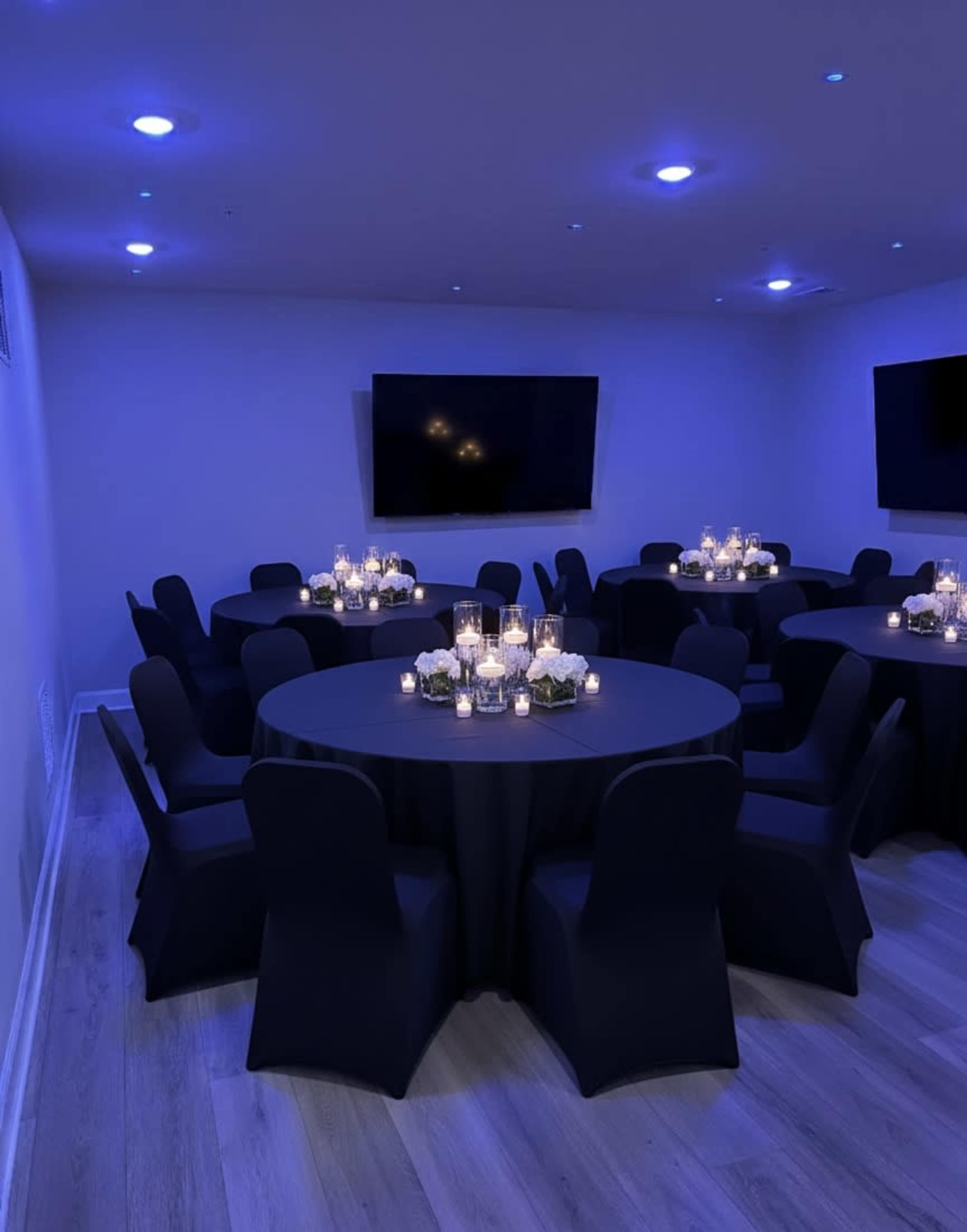 A dimly lit event room features several round tables covered with black tablecloths, each adorned with candles and floral arrangements.