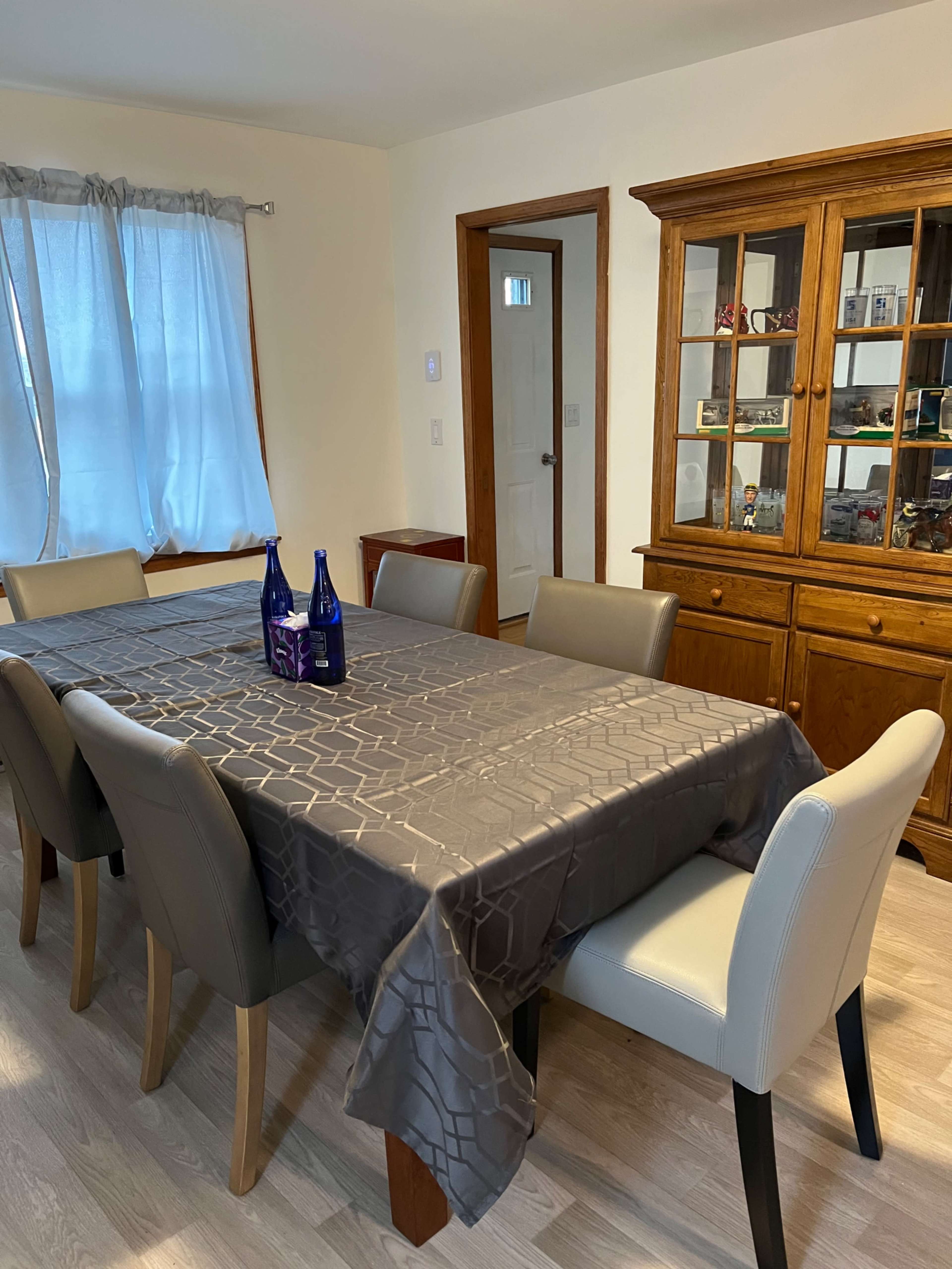 A dining table with a gray tablecloth and two blue bottles sits in a well-lit room with a side cabinet and a large window.