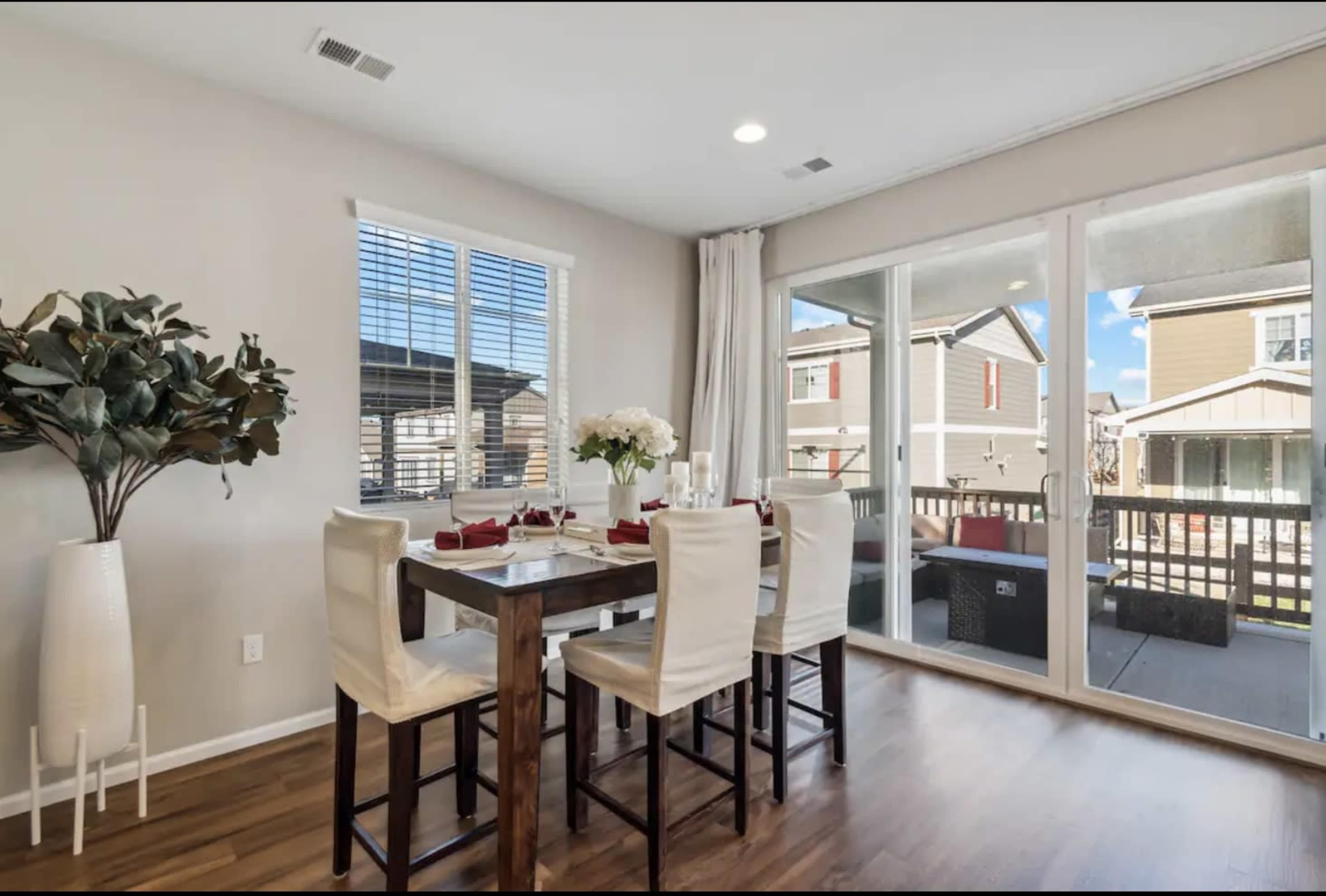 A dining area features a wooden table with white chairs, large windows allowing natural light, and a view of a patio.