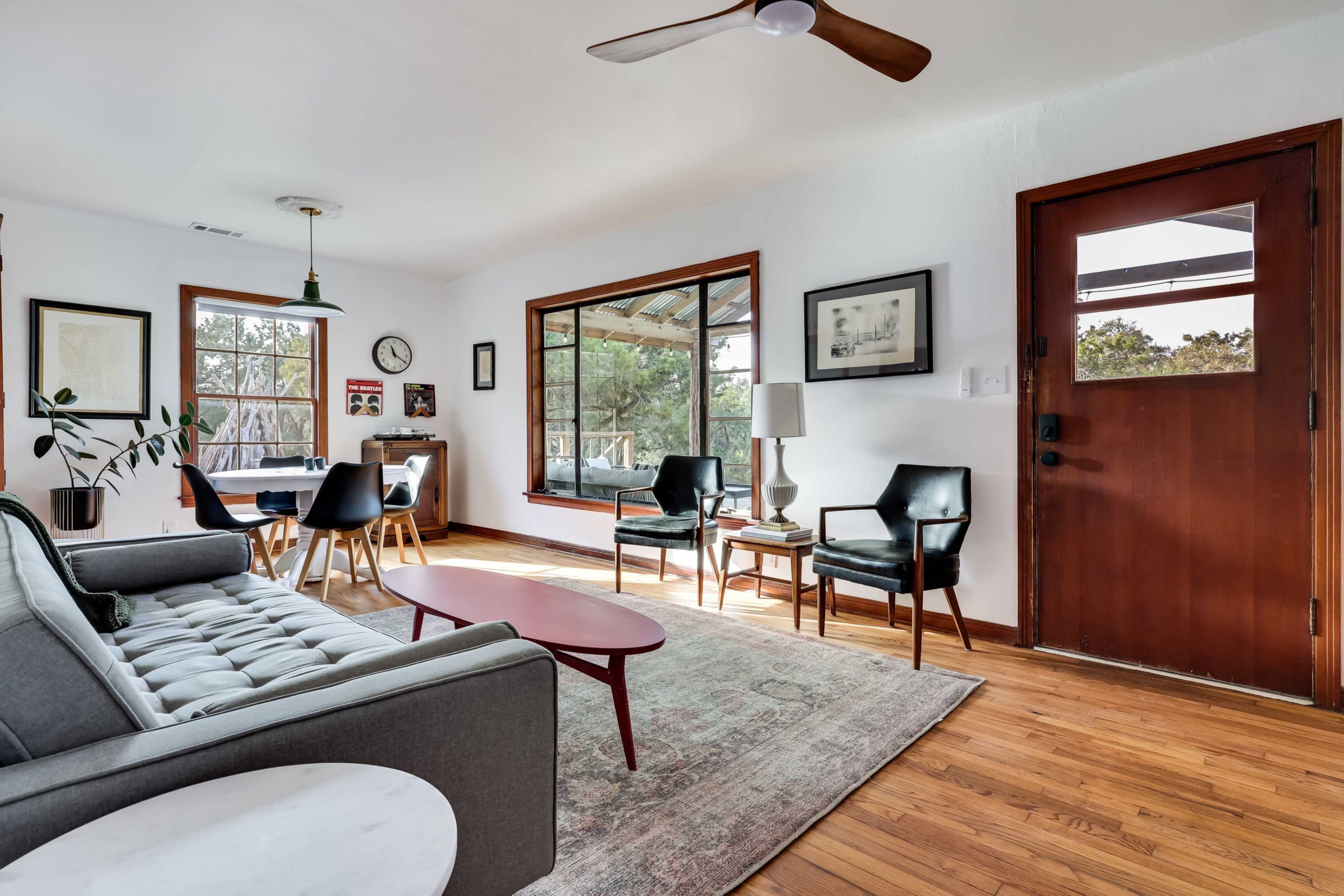 The image shows a modern living room with a gray sofa, wooden floor, a round coffee table, and a window overlooking a scenic outdoor view.