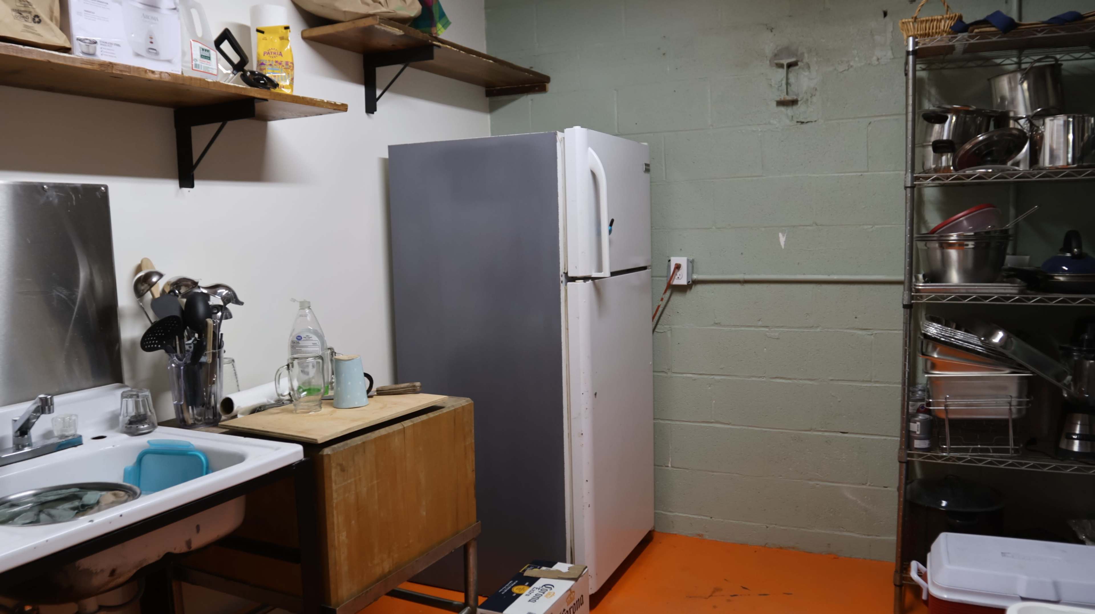 The image shows a small kitchen area with a sink, a wooden table, a grey refrigerator, and shelves containing various kitchen items.