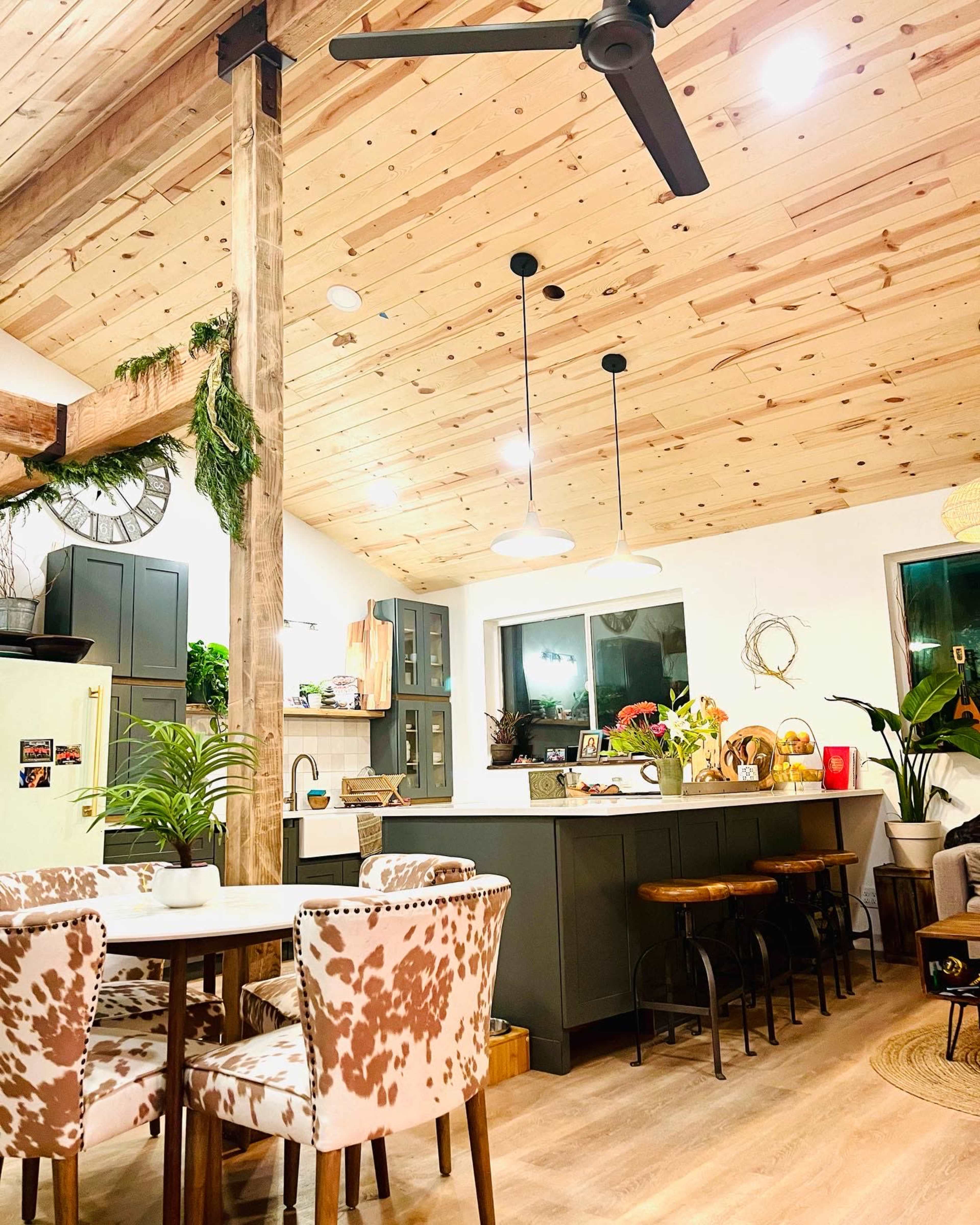 A modern kitchen and dining area with wooden beams, a light wooden ceiling, and a table surrounded by patterned chairs, adjacent to a kitchen island with bar stools.