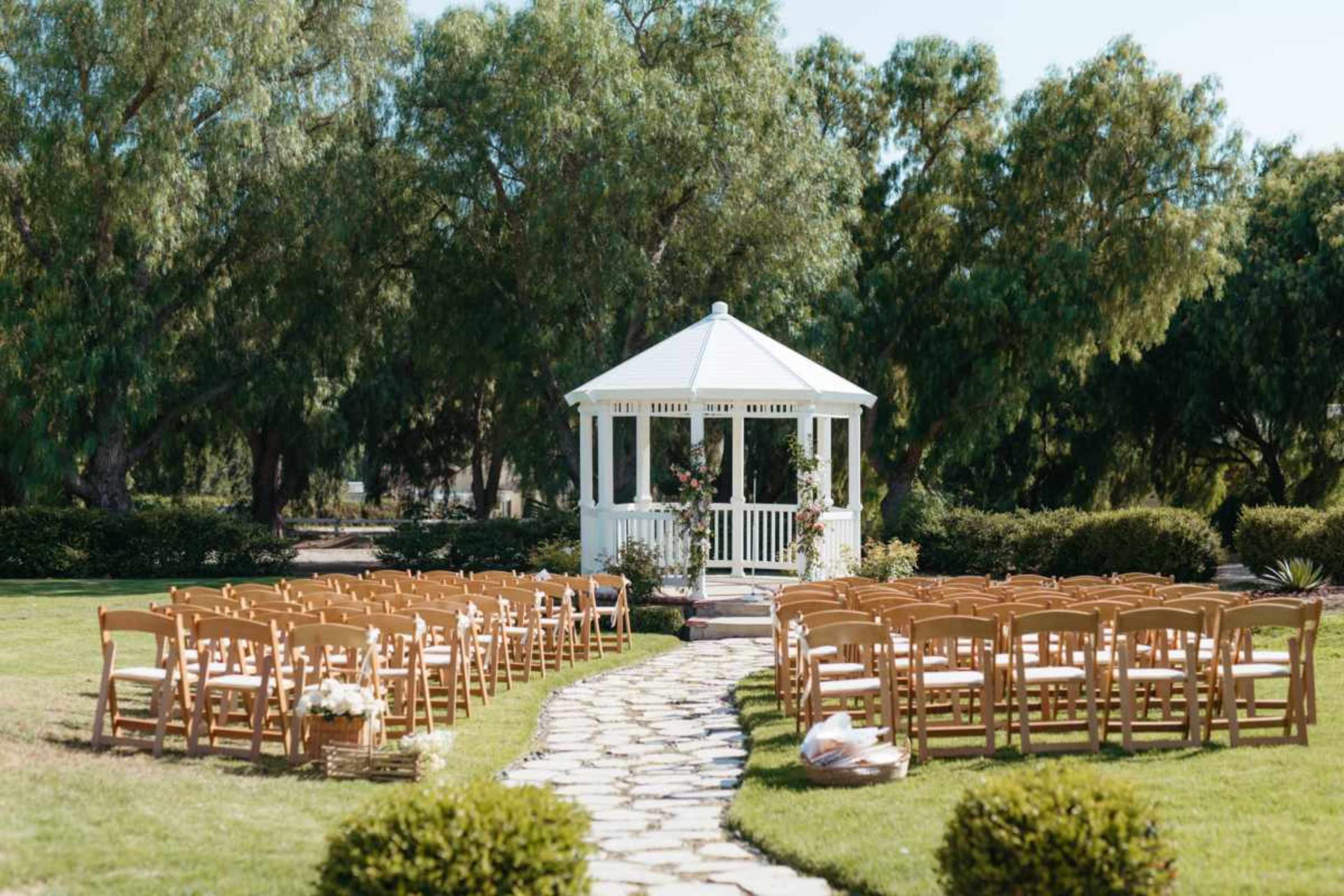 A white gazebo is surrounded by rows of wooden chairs set up on a stone pathway in a grassy outdoor space.